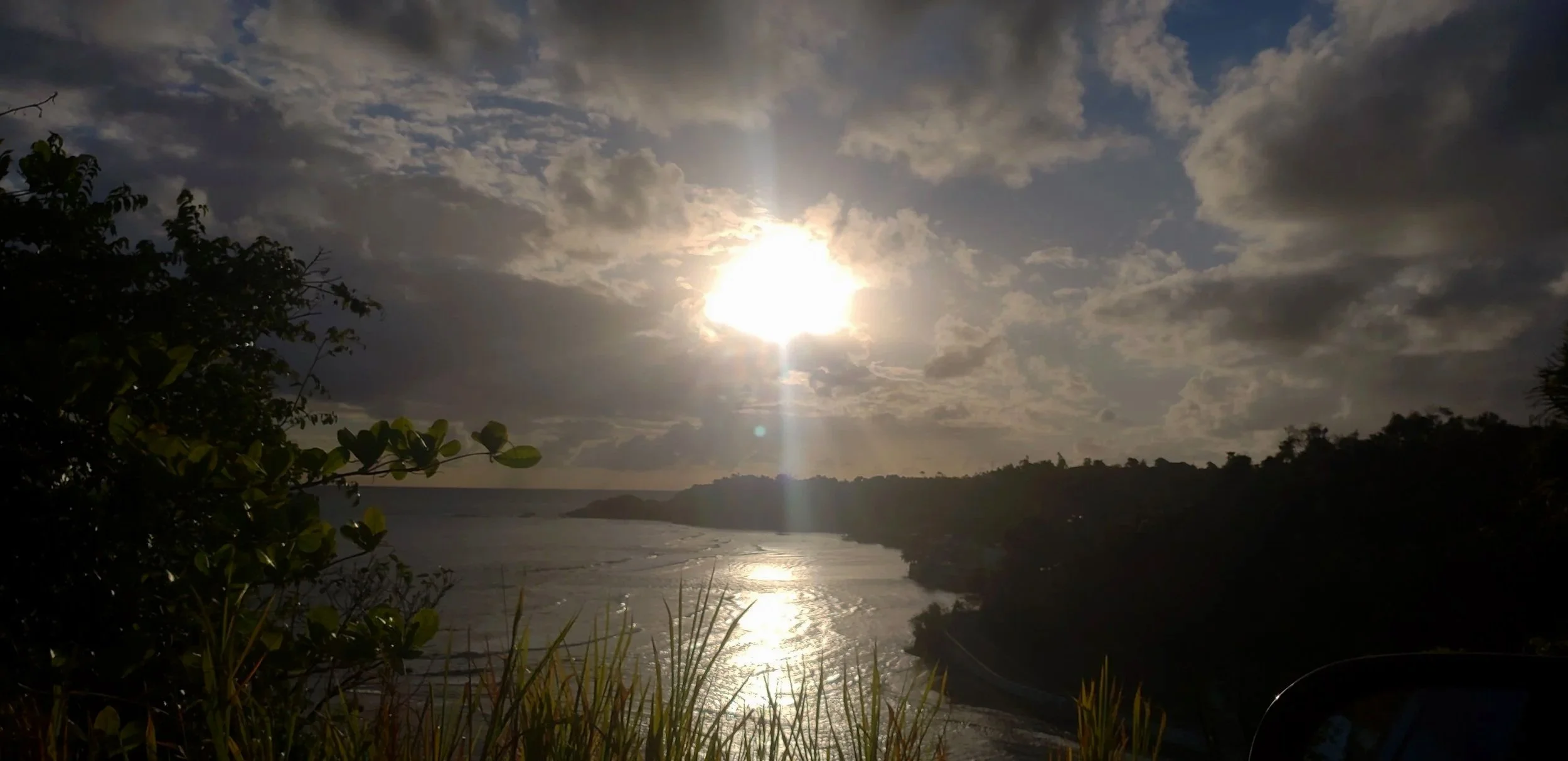 Sun partially obscured by clouds over a river with vegetation in the foreground and hills in the background.