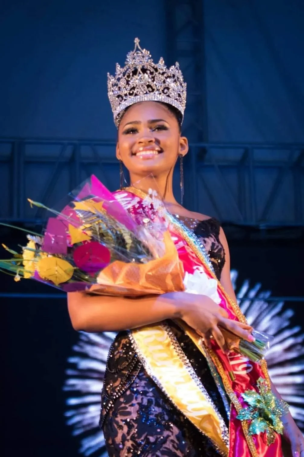Beauty pageant winner wearing a crown and sash, smiling, holding a bouquet of flowers, in a dark background with stage lighting.