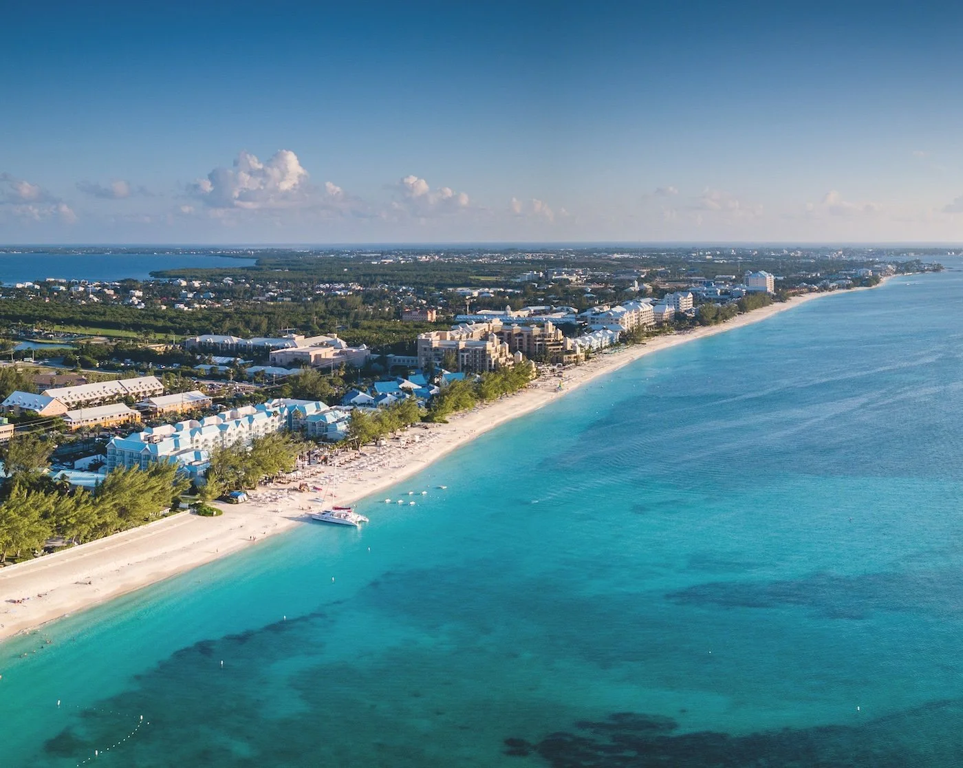 Aerial view of a coastal city with a sandy beach, blue water, and buildings lined along the shoreline, under a clear sky with clouds.