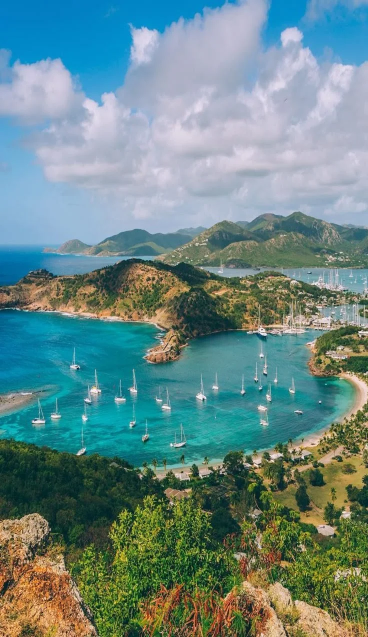 A coastal scene featuring a bay with sailboats, surrounded by green hills and mountains under a partly cloudy sky, with a marina visible in the distance.