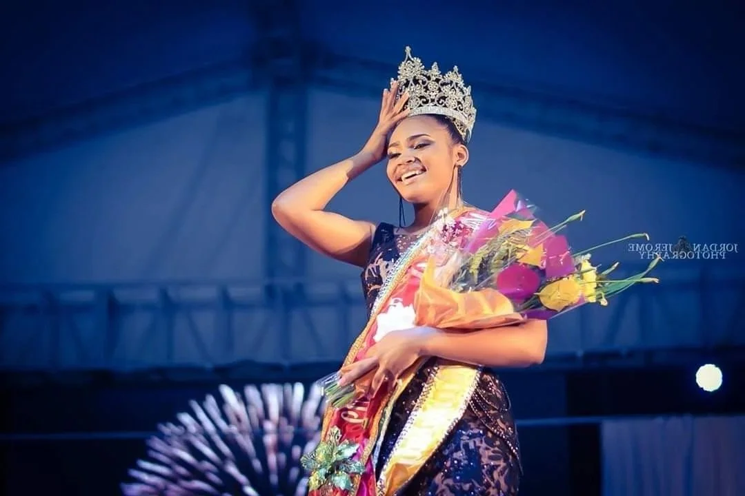A woman wearing a crown, holding a bouquet of flowers, smiling, and dressed in a sash and evening gown on stage.