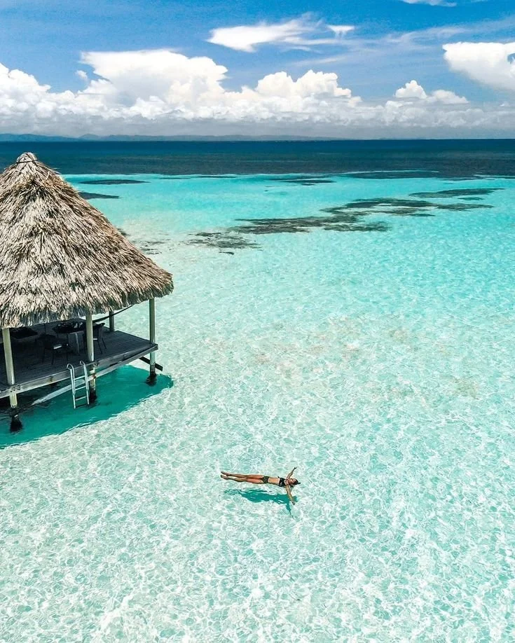 A woman lying on shallow, clear, turquoise water near a thatched-roof hut at a tropical beach.