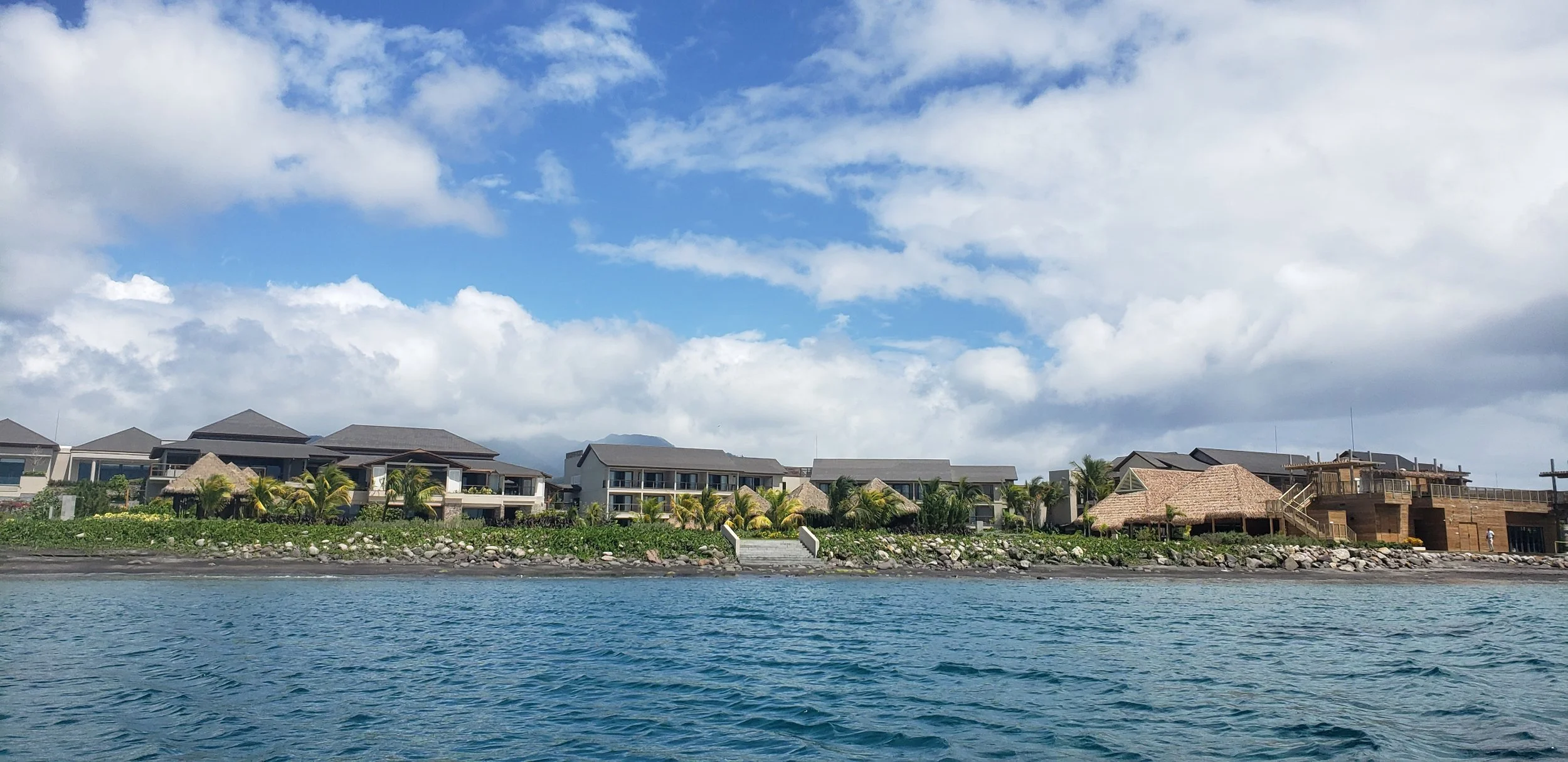 Ocean view with water in the foreground, a shoreline with rocks and vegetation, and modern buildings with thatched roofs and palm trees in the background under a partly cloudy sky.