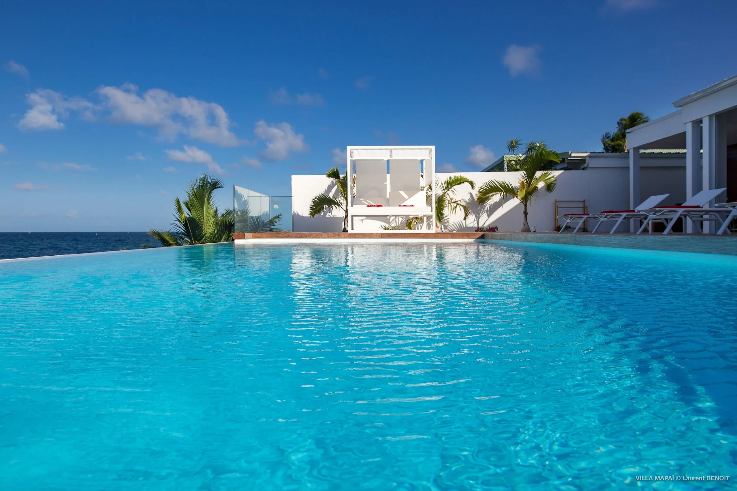 A view of a swimming pool overlooking the ocean under a blue sky with white clouds. There are lounge chairs and tropical plants near the pool.
