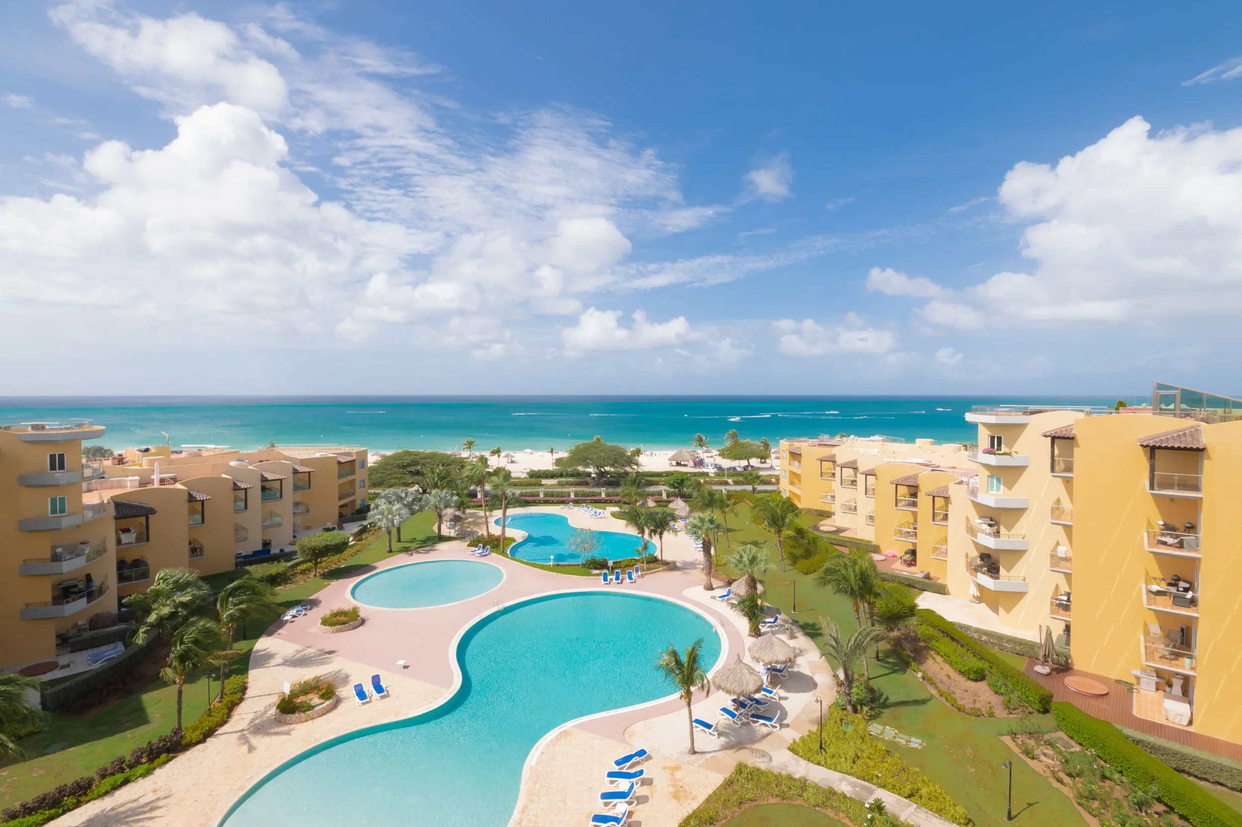 View of a beachside resort with a large swimming pool, surrounded by yellow buildings with balconies, and a tropical setting with palm trees. The ocean and sky are visible in the background.