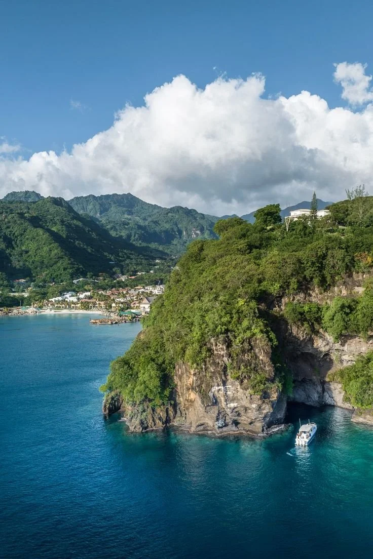 A coastal landscape with green mountainous cliffs, a blue ocean, and a boat entering a small cave in the rocks.