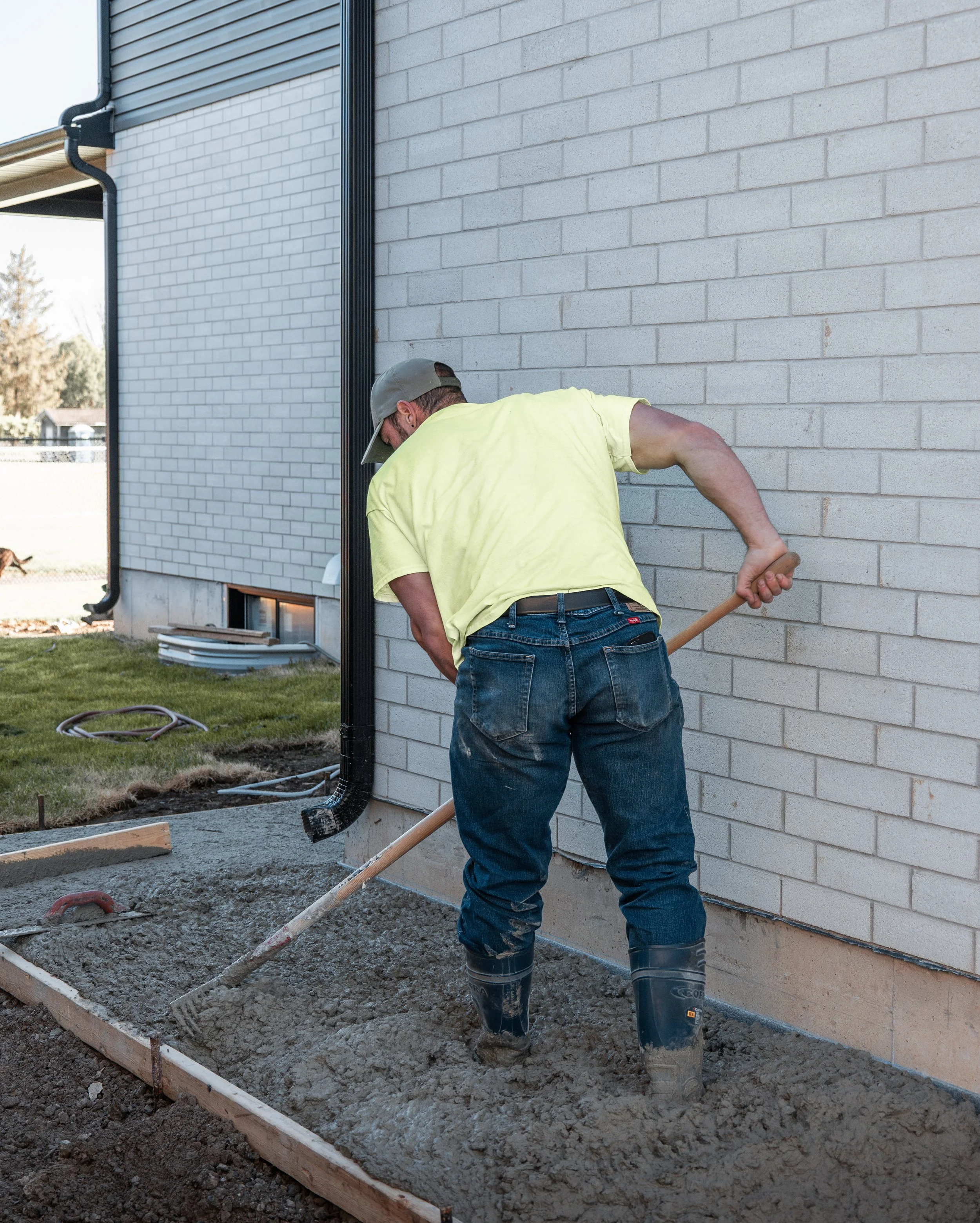 A man wearing a gray cap, yellow t-shirt, and jeans is leveling wet concrete with a rake in a garden bed next to a house with white brick siding.