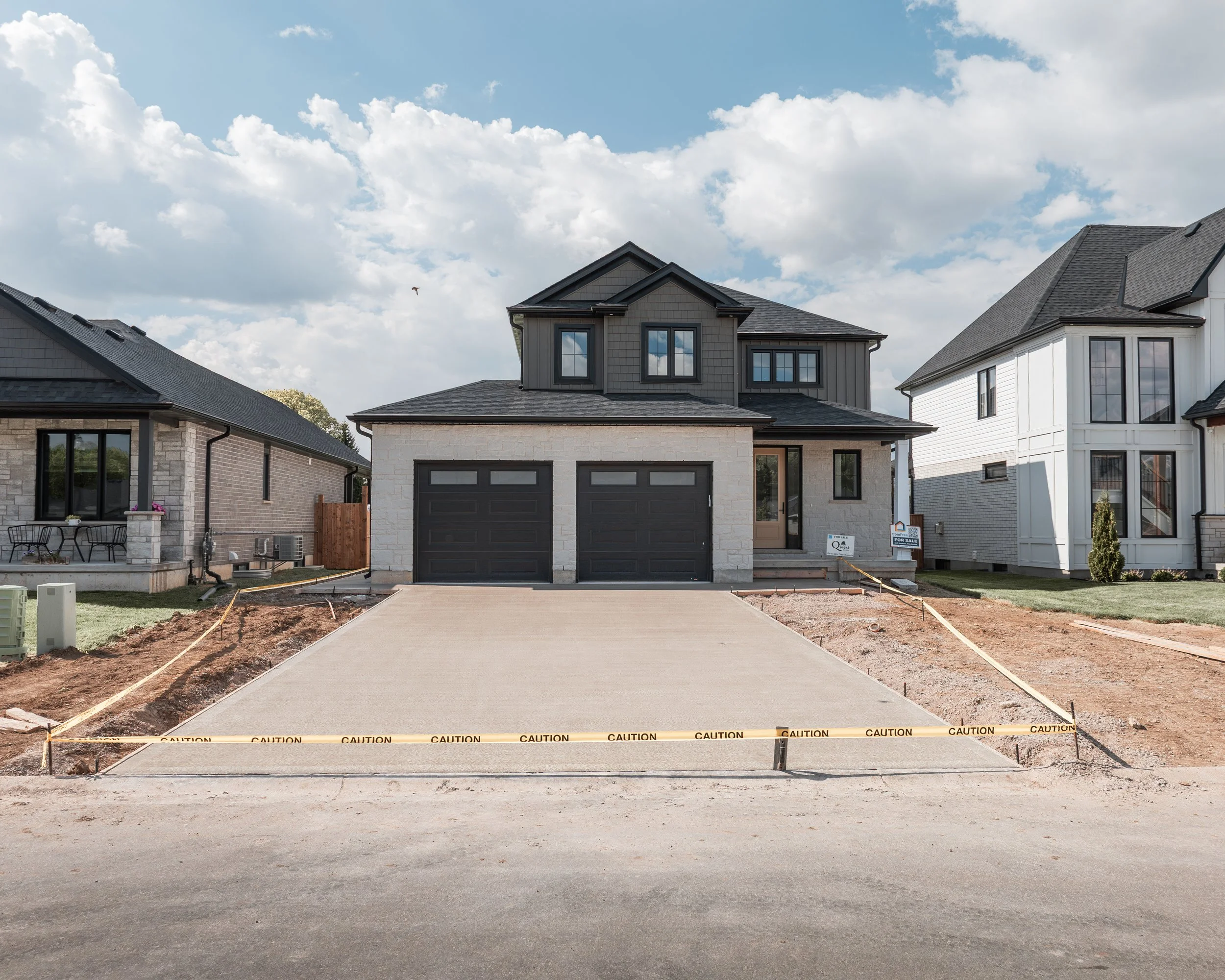 Newly constructed two-story house with a black garage door, gray exterior, and a concrete driveway under construction. Caution tape surrounds the front yard, and neighboring houses are visible on either side under a partly cloudy sky.