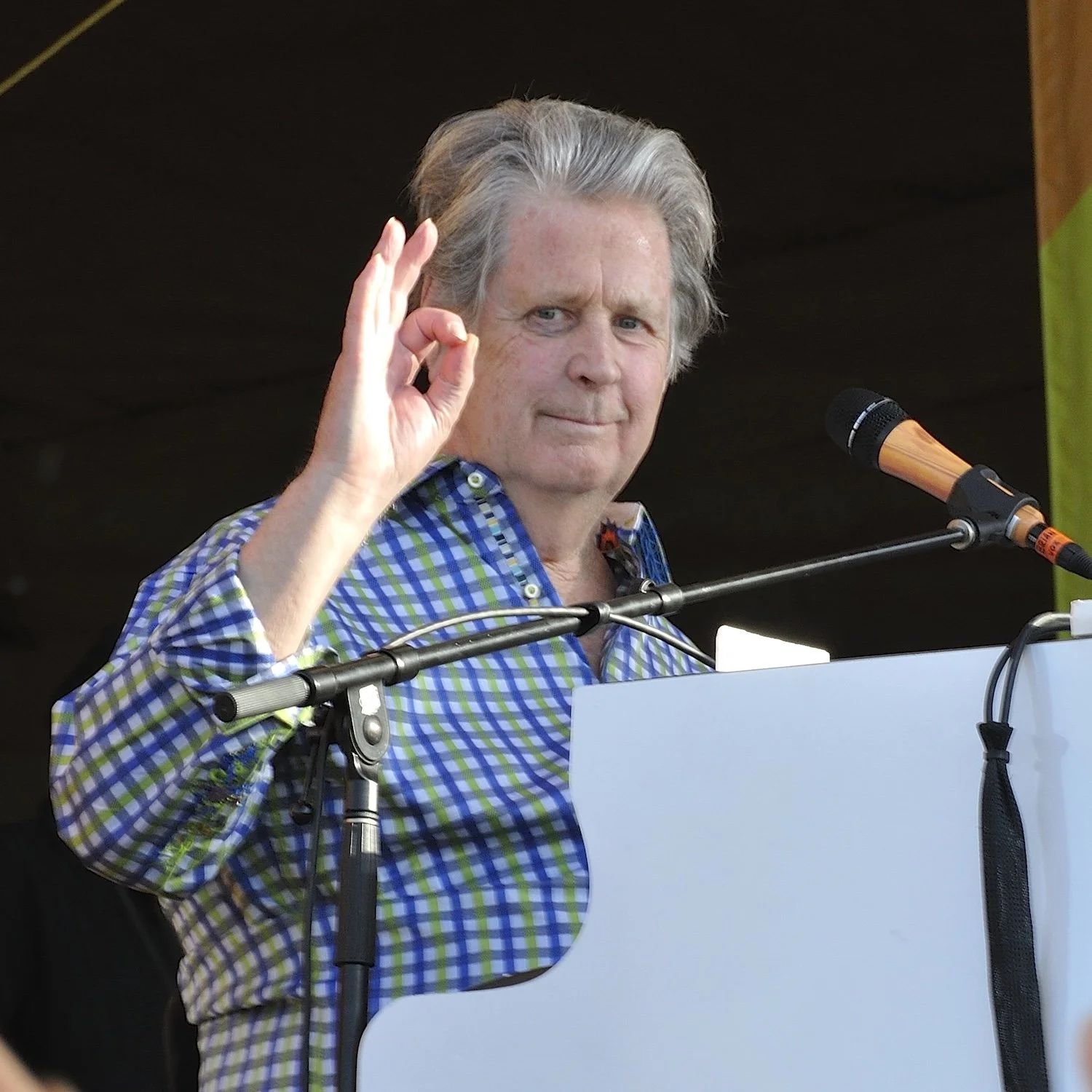 Older man with gray hair at a podium, making an OK gesture with his hand, speaking into a microphone while wearing a blue and green checkered shirt.