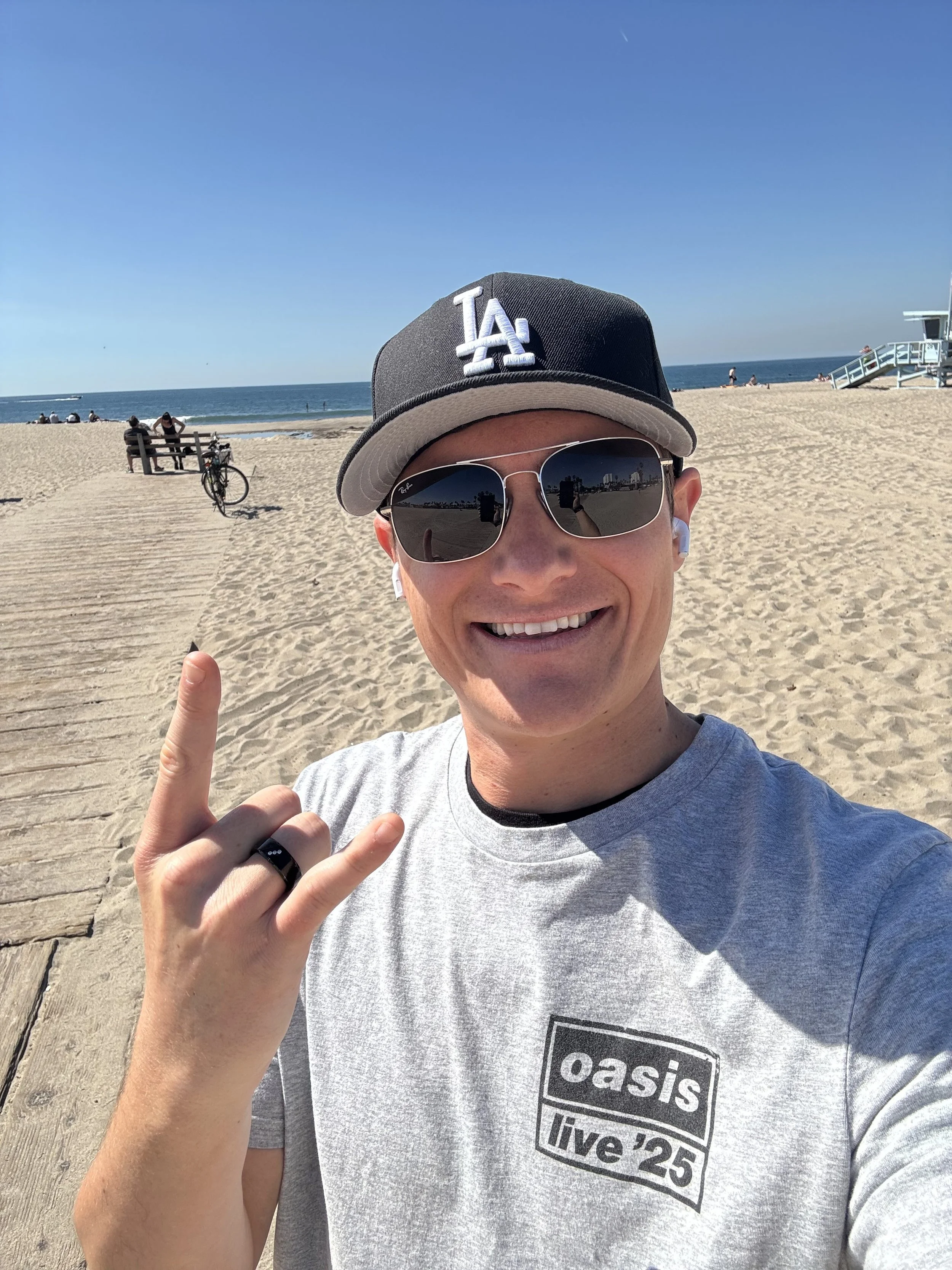 Founder at the beach wearing a Los Angeles Dodgers cap, sunglasses, and a gray Oasis T-shirt, smiling and making a rock on hand gesture, with a bicycle, benches, lifeguard tower, and the ocean in the background.