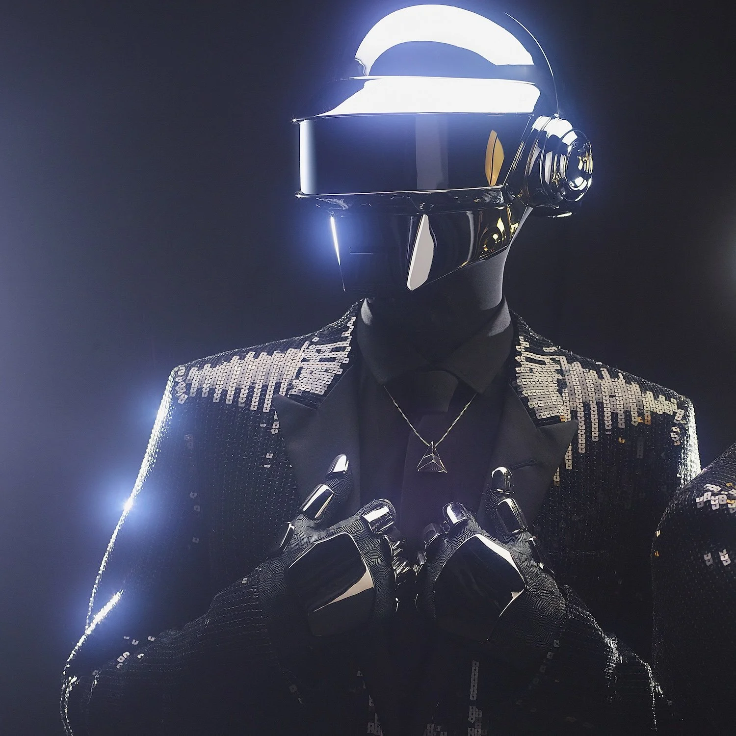 A person wearing a shiny black helmet with a reflective visor, dressed in a sequined black jacket with silver decorations. They are holding their hands over their chest with metallic gloves, and are wearing a necklace with a geometric pendant. The background is dark with subtle lighting highlights.