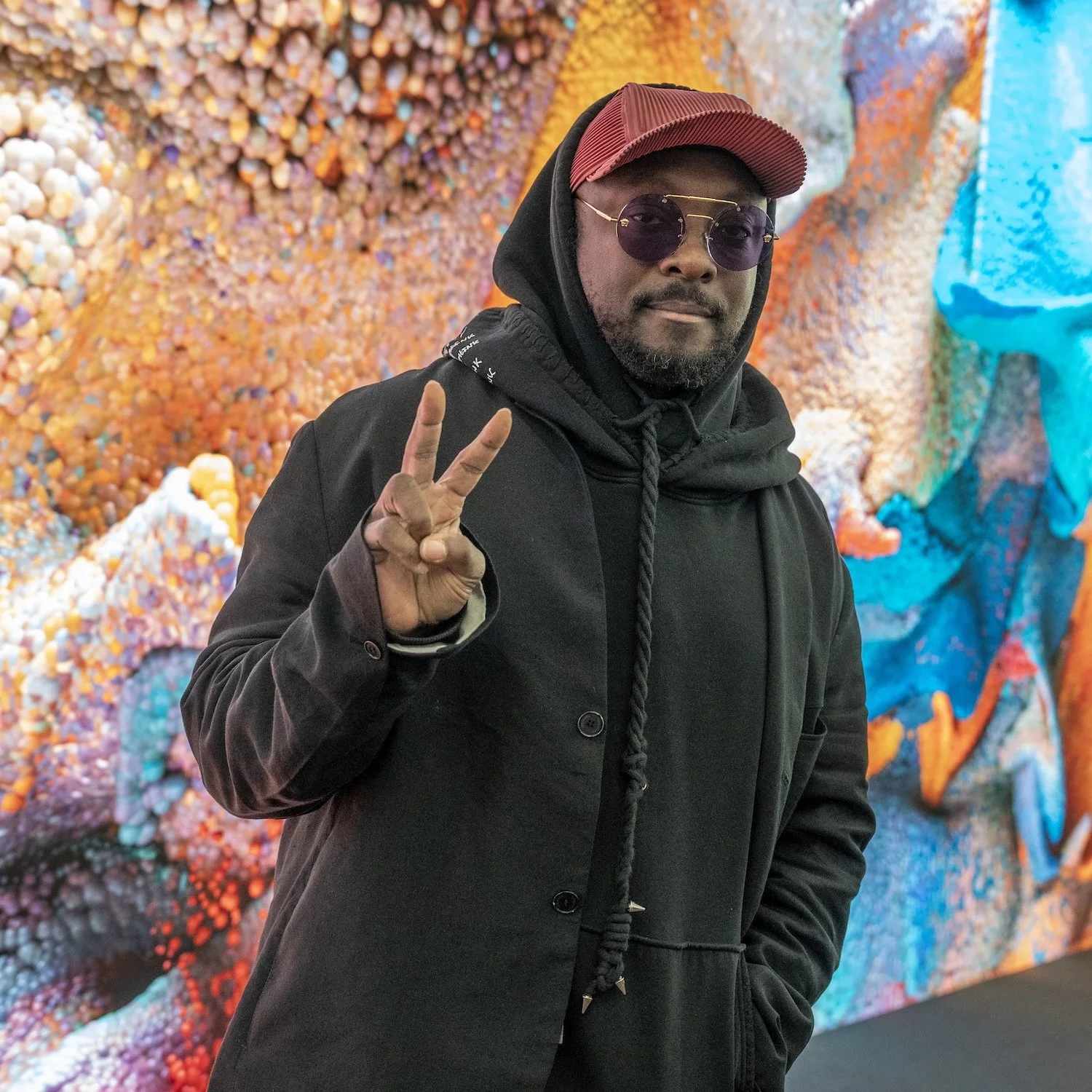 A man with dark skin wearing a red cap, purple sunglasses, a black hoodie, and a black jacket making a peace sign with his right hand, standing in front of a colorful abstract background.