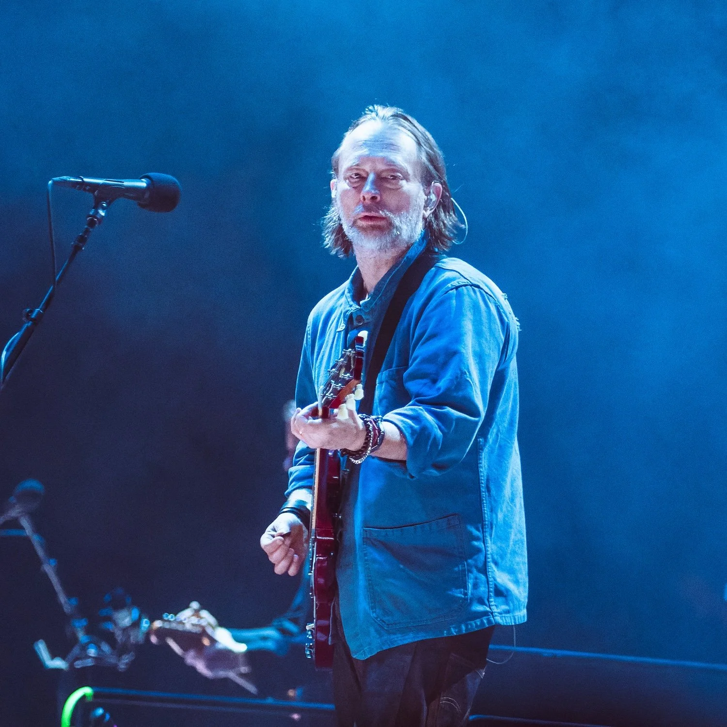 A male musician with long hair and beard playing an electric guitar on stage, illuminated by blue stage lighting, with a microphone nearby.