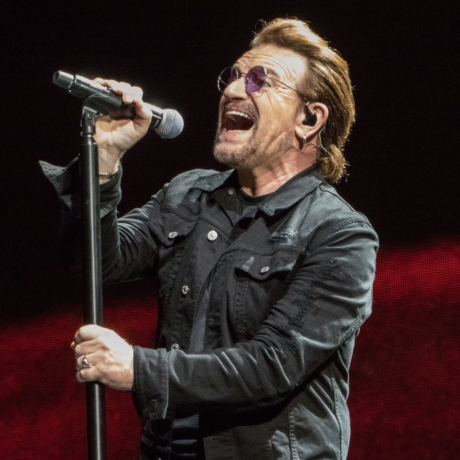 Male singer passionately performing on stage with microphone, wearing sunglasses, gray jacket, and earrings.