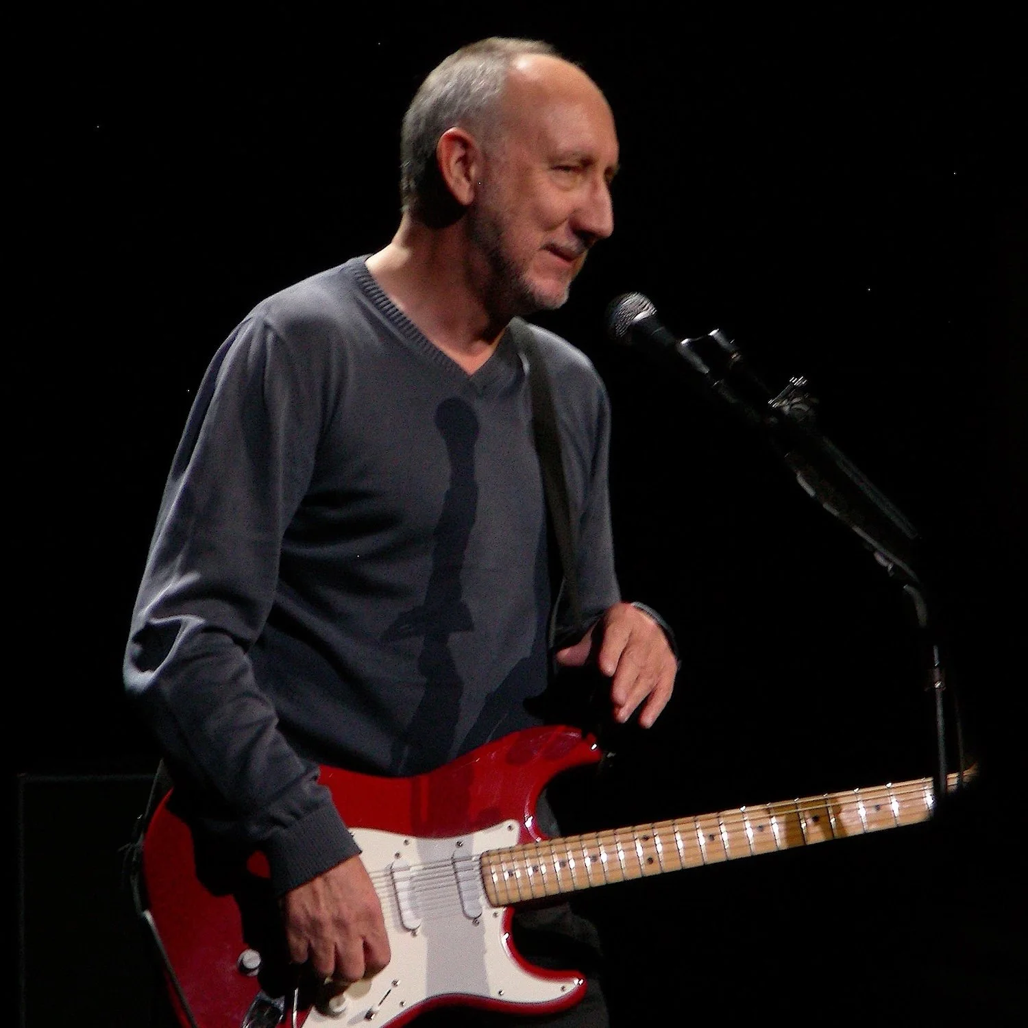 An older man with gray hair and a beard, wearing a gray long-sleeve shirt, stands on stage with a red and white electric guitar. He is near a microphone, smiling softly, with a dark background.