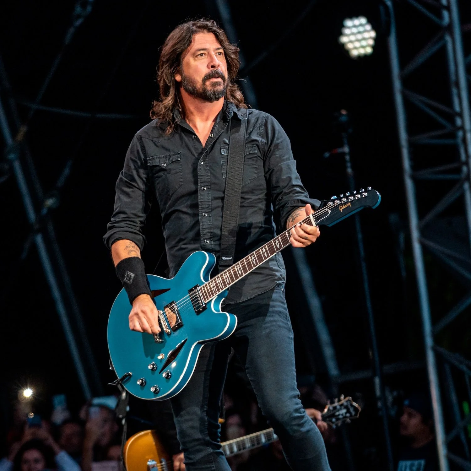 A male guitarist with long dark hair and a beard, wearing a black shirt and black pants, playing a blue electric guitar on stage during a concert.