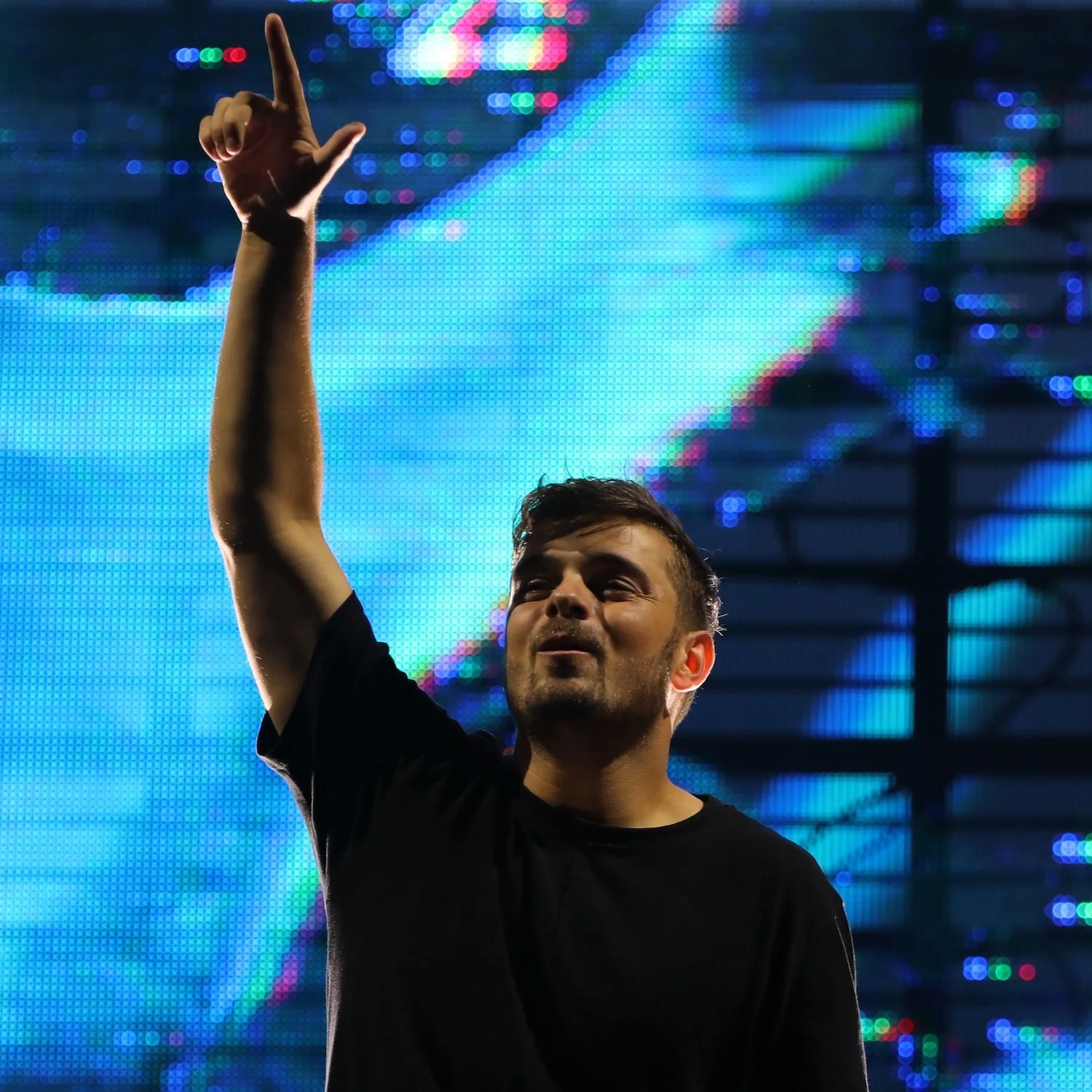 A man in a black shirt raising his right hand with his thumb and pinky finger extended, standing in front of a digital LED screen with colorful, blurred lights.