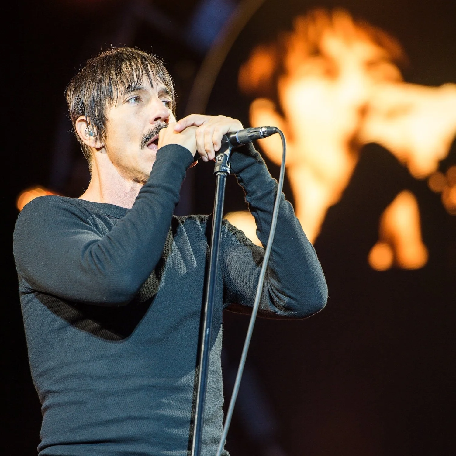 A male singer with dark hair and a mustache singing into a microphone on stage, with a large screen displaying an abstract image behind him.
