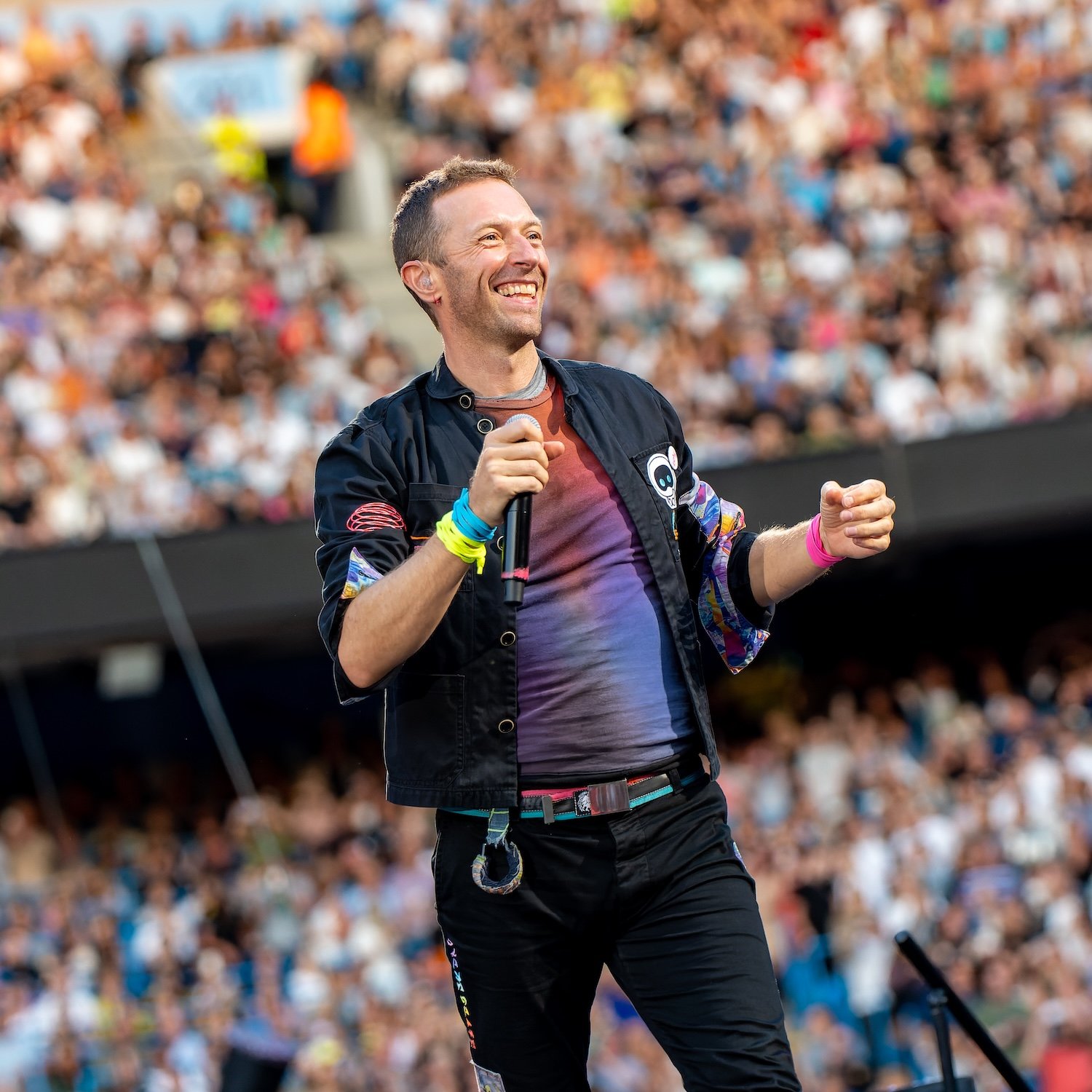 A man with short dark hair and a beard smiling while holding a microphone at a crowded outdoor event.