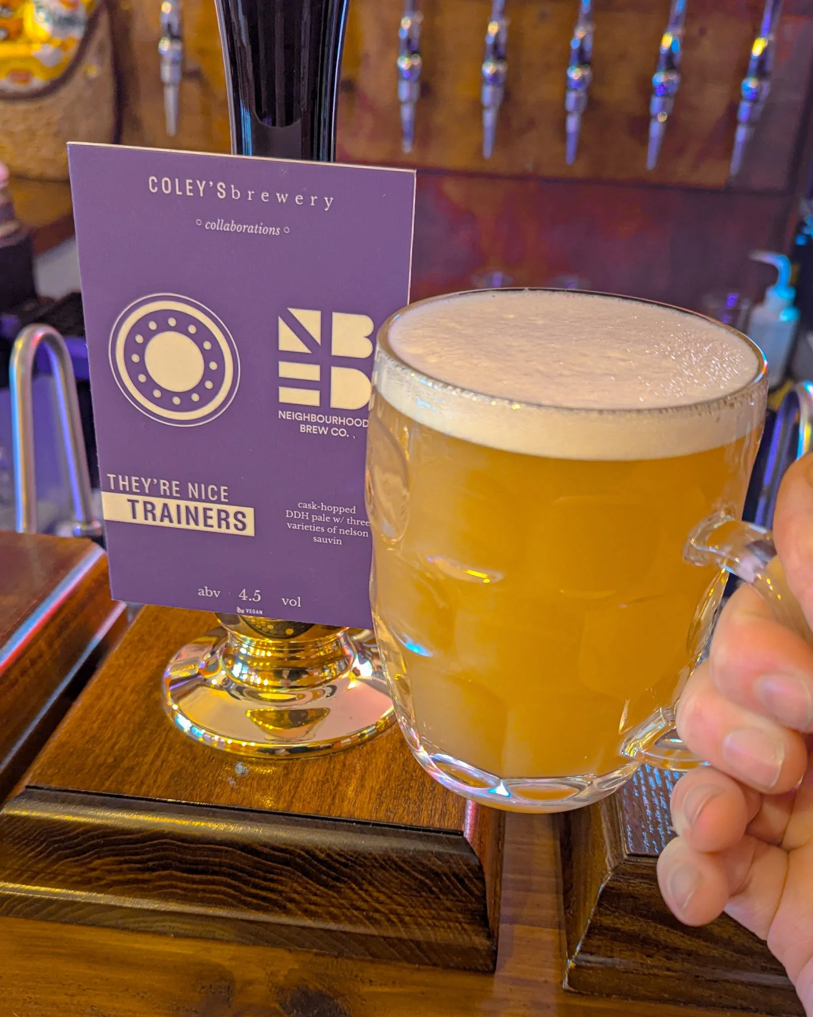 A glass mug of pale beer with a foamy head held by a person's hand, placed on a wooden table with a drink menu and taps in the background.
