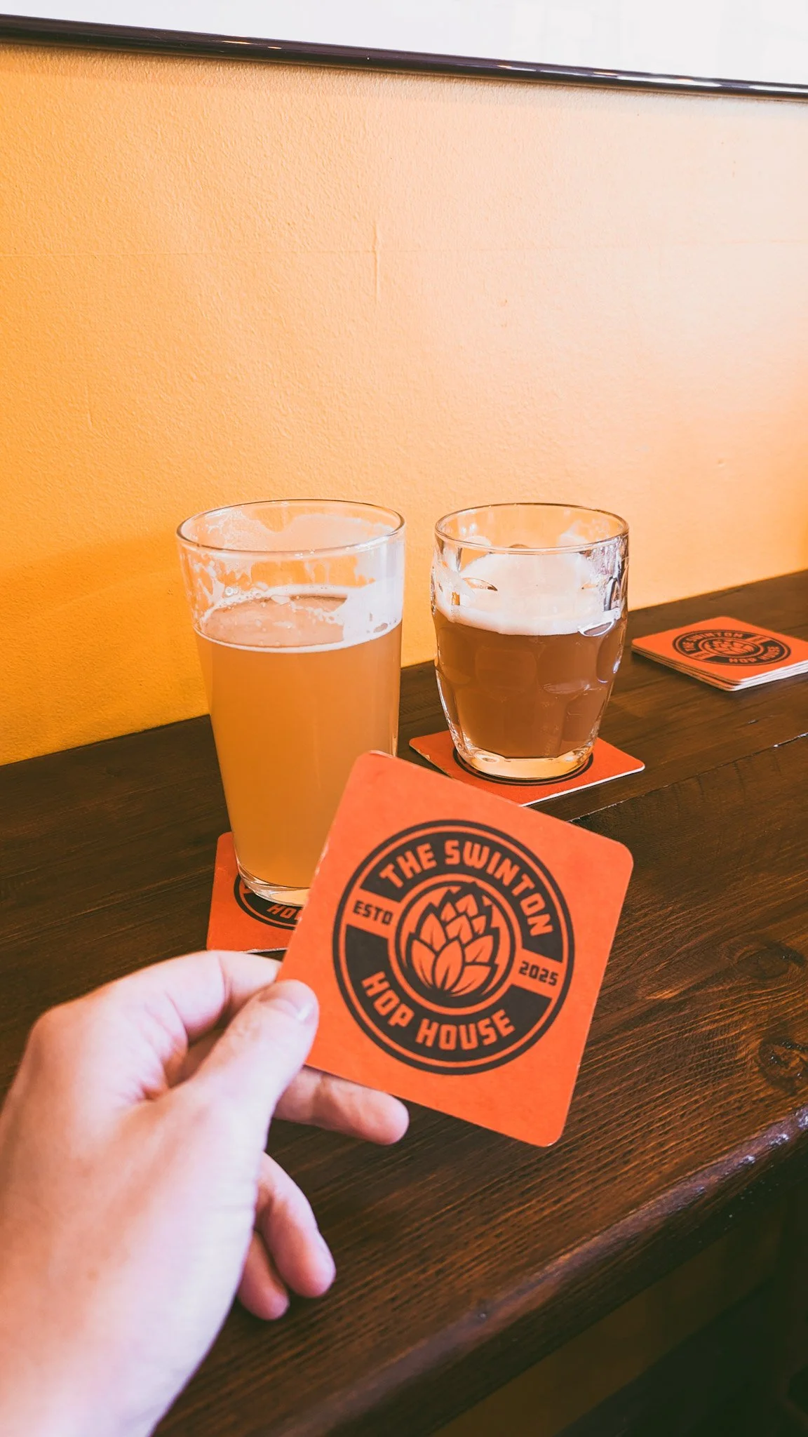 Two glasses of beer, one light and one dark, on a wooden bar counter with coasters. A person is holding an orange coaster with a hop cone logo and the text 'The Swinton Hop House, ESTD 2025'.