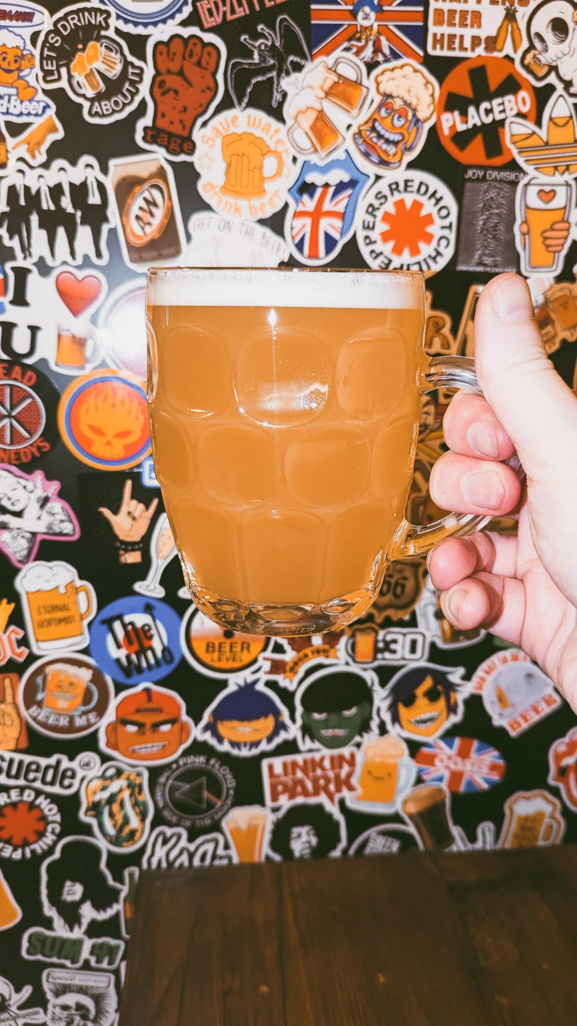 A hand holding a clear glass mug filled with a frothy, light-colored beer. The background features a wall decorated with various colorful stickers, many related to beer, music, and pop culture.