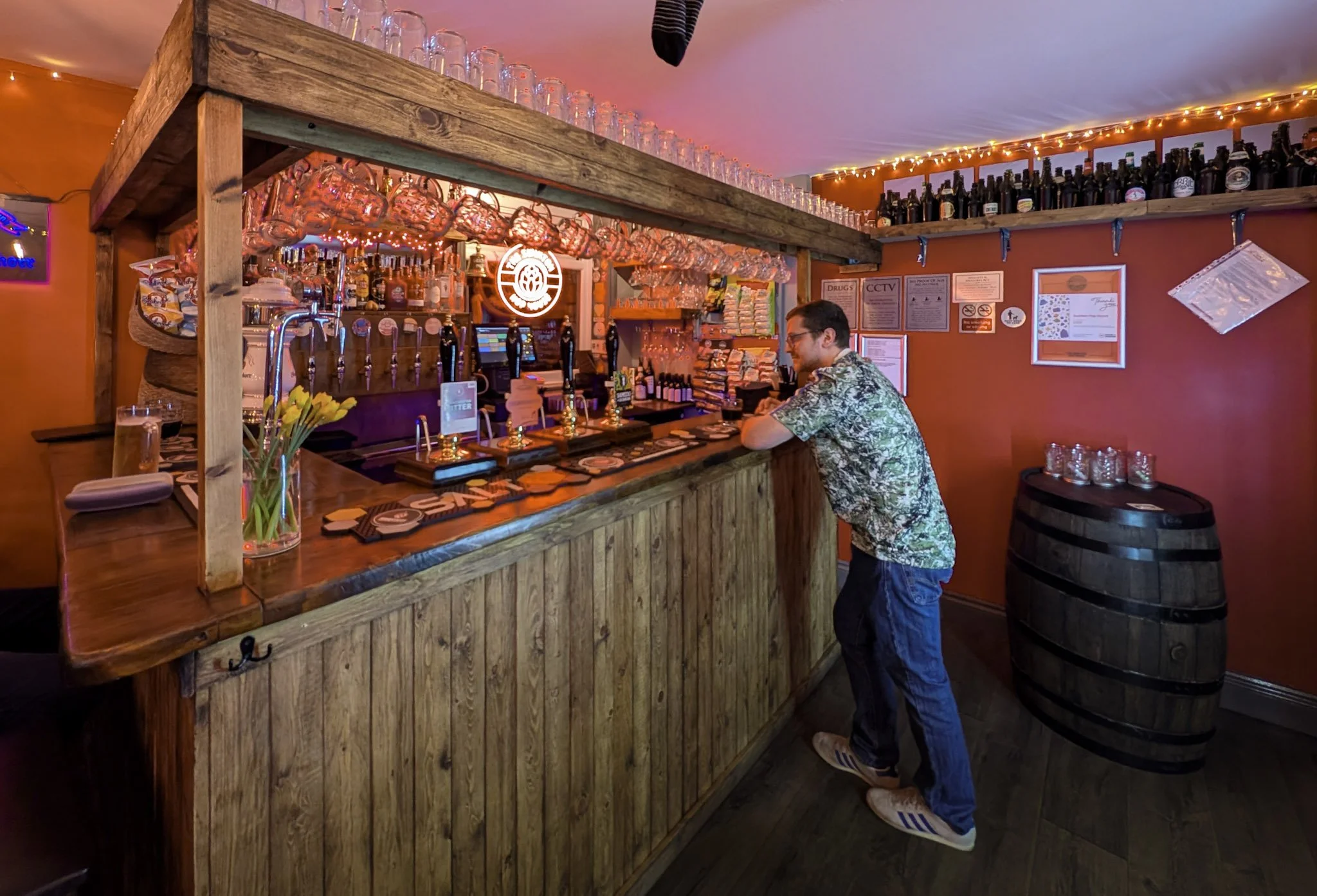 A man standing at a wooden bar counter in a cozy bar or pub, with an assortment of beer taps and glasses behind it. The bar area has warm lighting, with bottles and glasses on shelves and a neon sign on the wall. The man is wearing glasses, a patterned shirt, jeans, and sneakers.