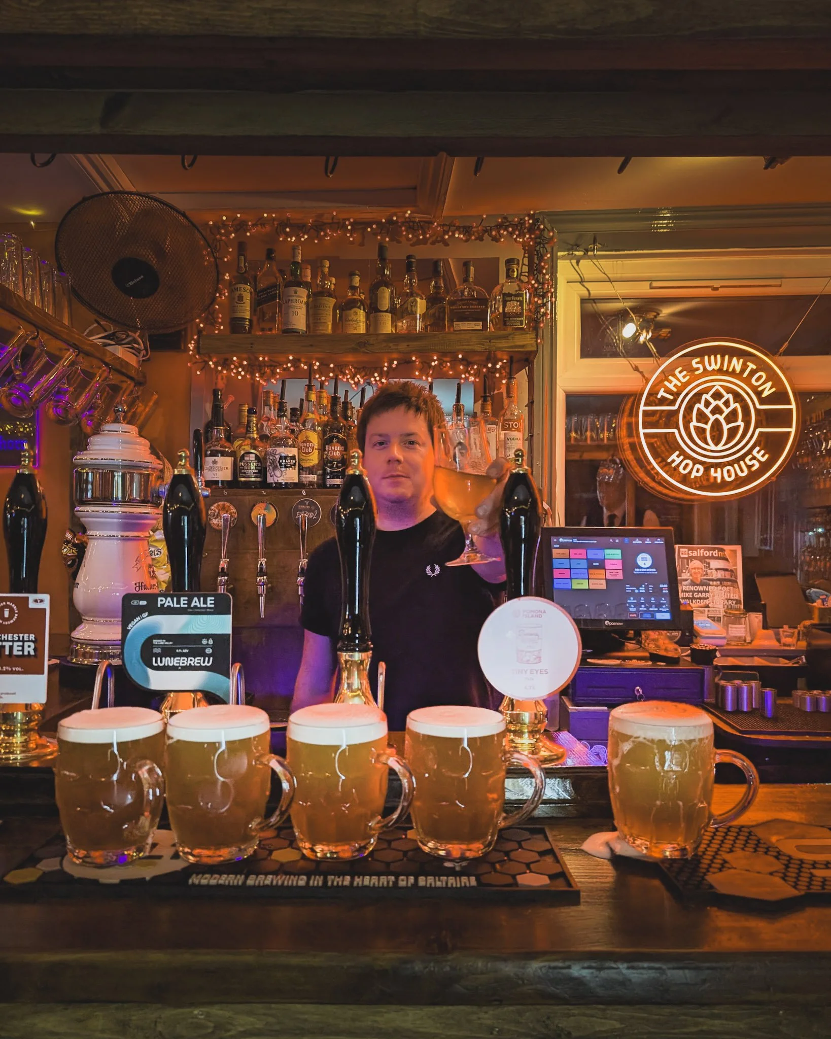 Bartender pouring beer at a bar with five beer mugs and a row of beer taps behind him, bottles of alcohol on shelves, neon sign that says 'The Swinton Hop House,' and warm ambient lighting.