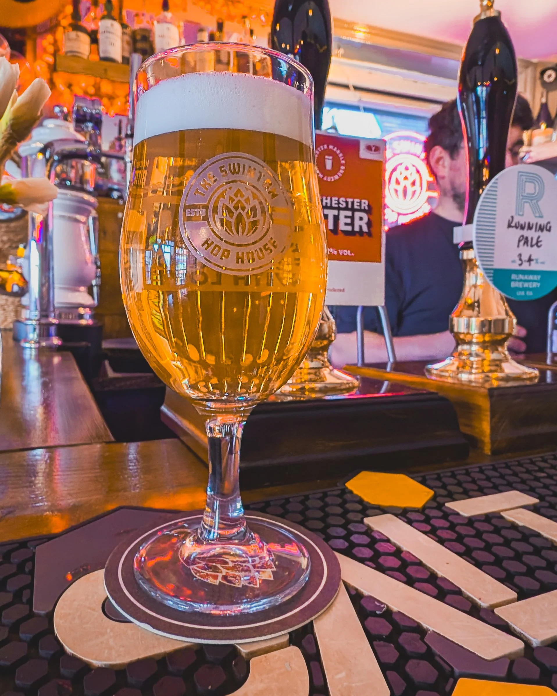 A glass of beer with a foam head on a bar table, with bar decorations, bottles, and neon signs in the background.