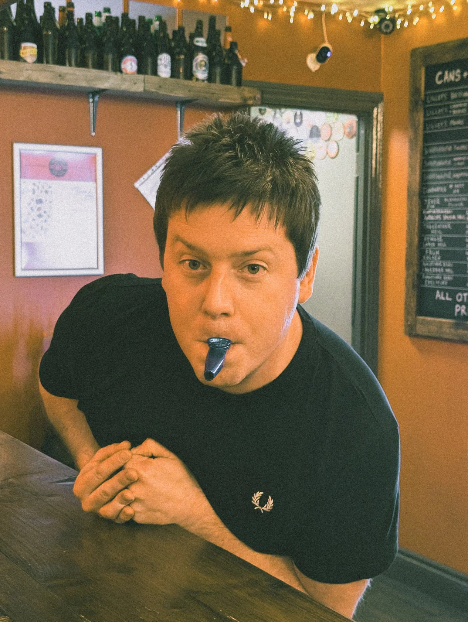 A man with short dark hair leaning over a wooden table, holding a party horn in his mouth, inside a cosy bar with orange walls, string lights, and a chalkboard menu.
