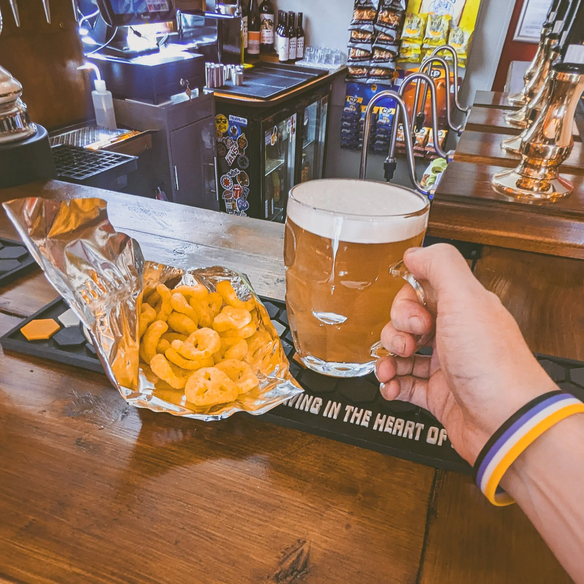 A hand holding a glass of beer next to a bag of cheese snacks on a bar counter.