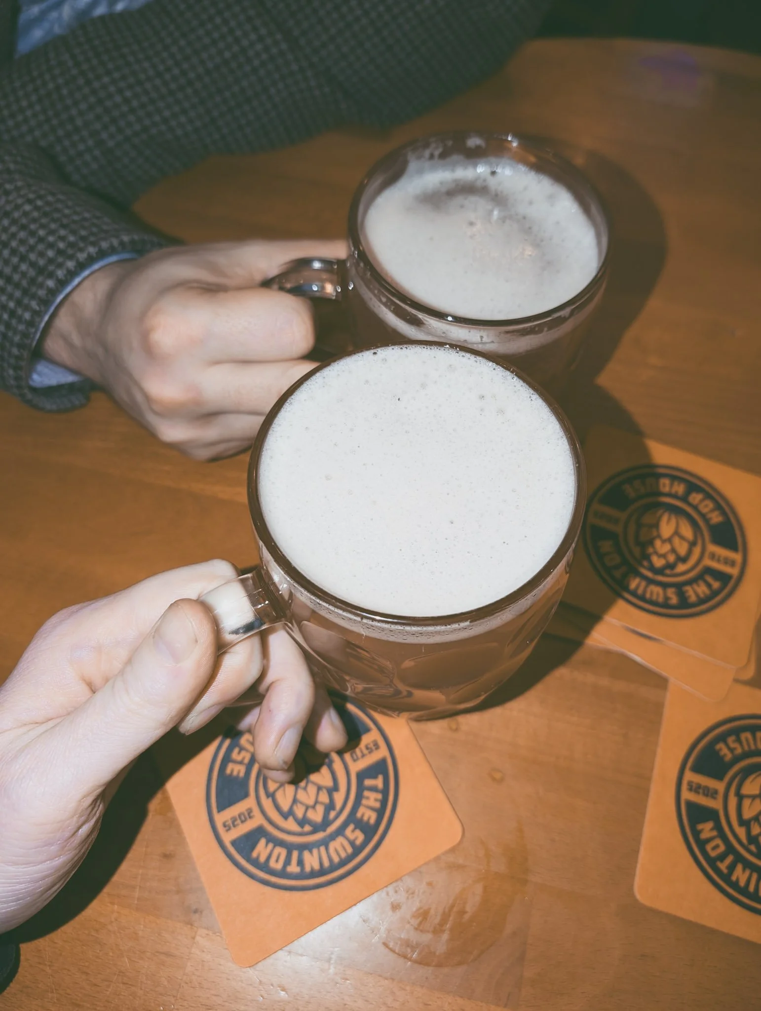 Two people clinking glasses of beer at a wooden table with promotional coasters from The Swinton Hop House.