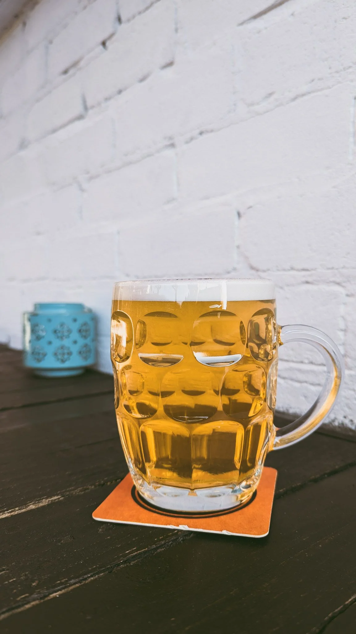 A clear glass mug filled with golden beer, sitting on an orange coaster on a dark wooden table with a white brick wall in the background.