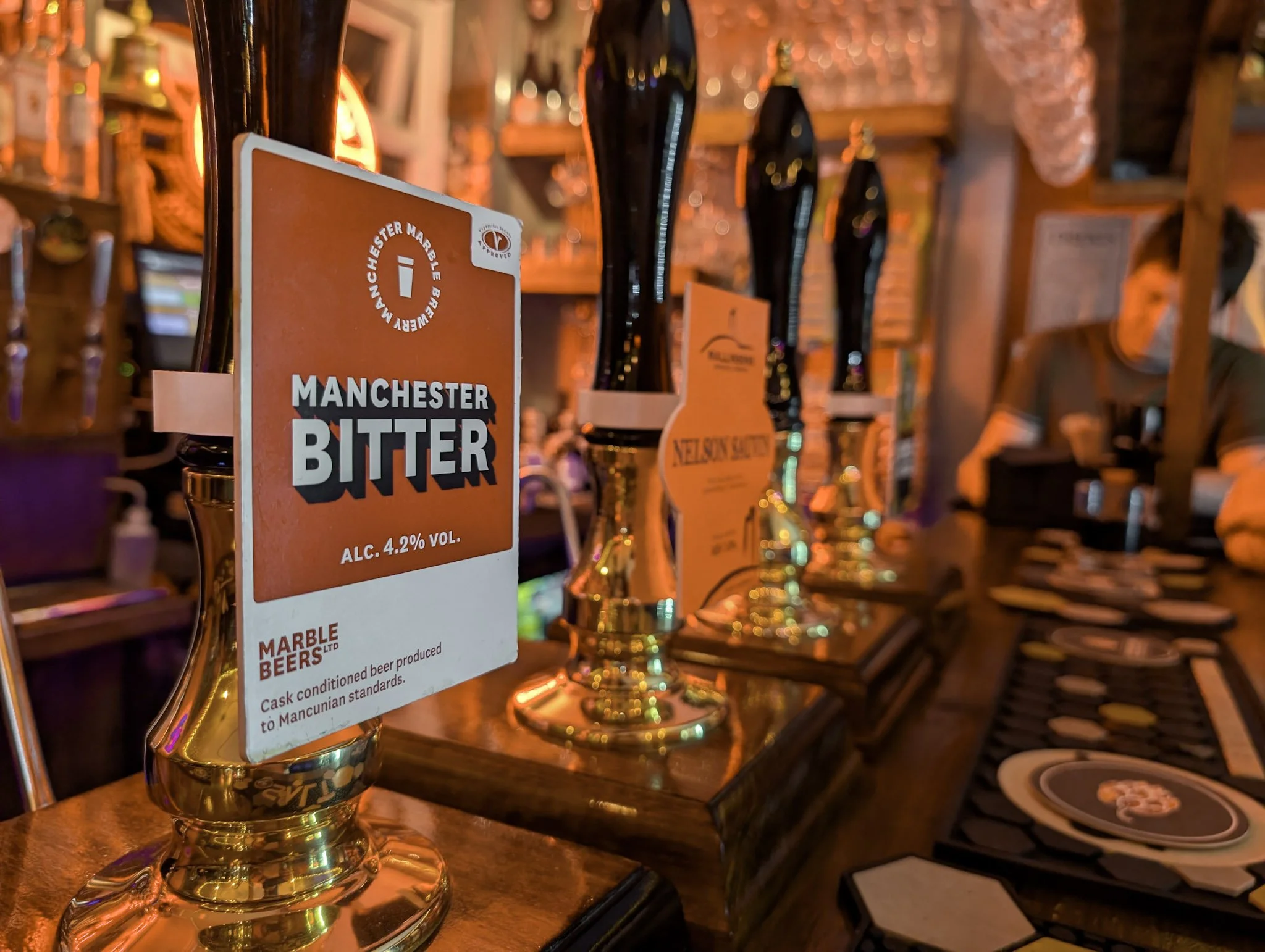 Bar scene with beer taps and a label for Manchester Bitter beer with 4.2% alcohol by volume, warm lighting, and a person in the background.