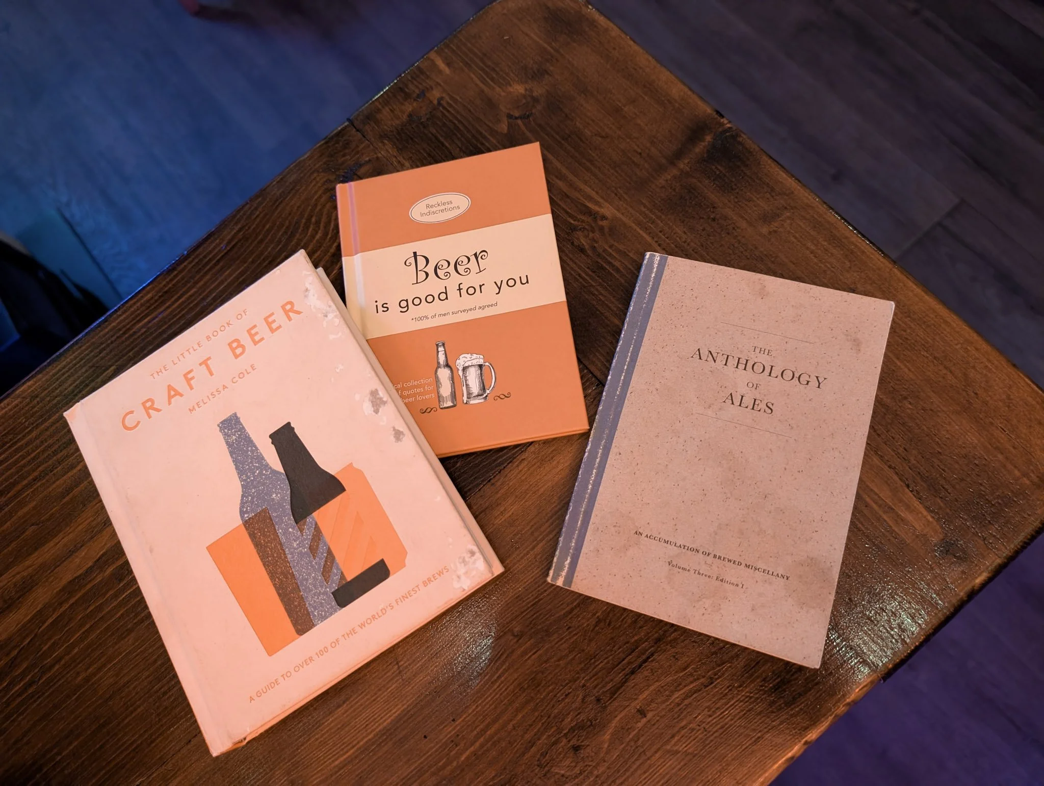 Three books about beer on a wooden table. The titles are 'The Little Book of Craft Beer' by Melissa Cole, 'Beer is Good for You', and 'The Anthology of Ales'.