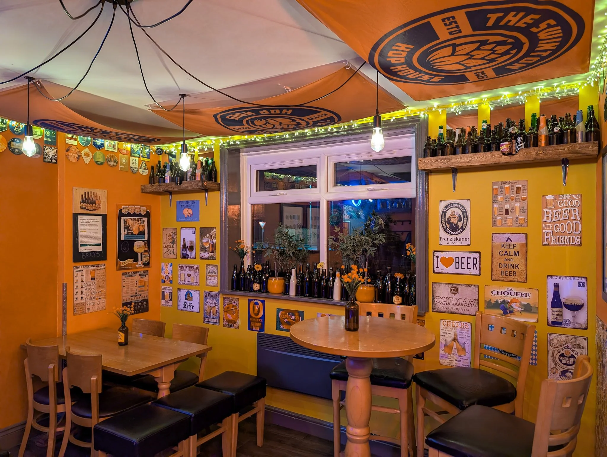 Decorated cosy bar with yellow walls, beer posters, shelves of bottles, tables with chairs, flowers in vases, and ceiling banners.