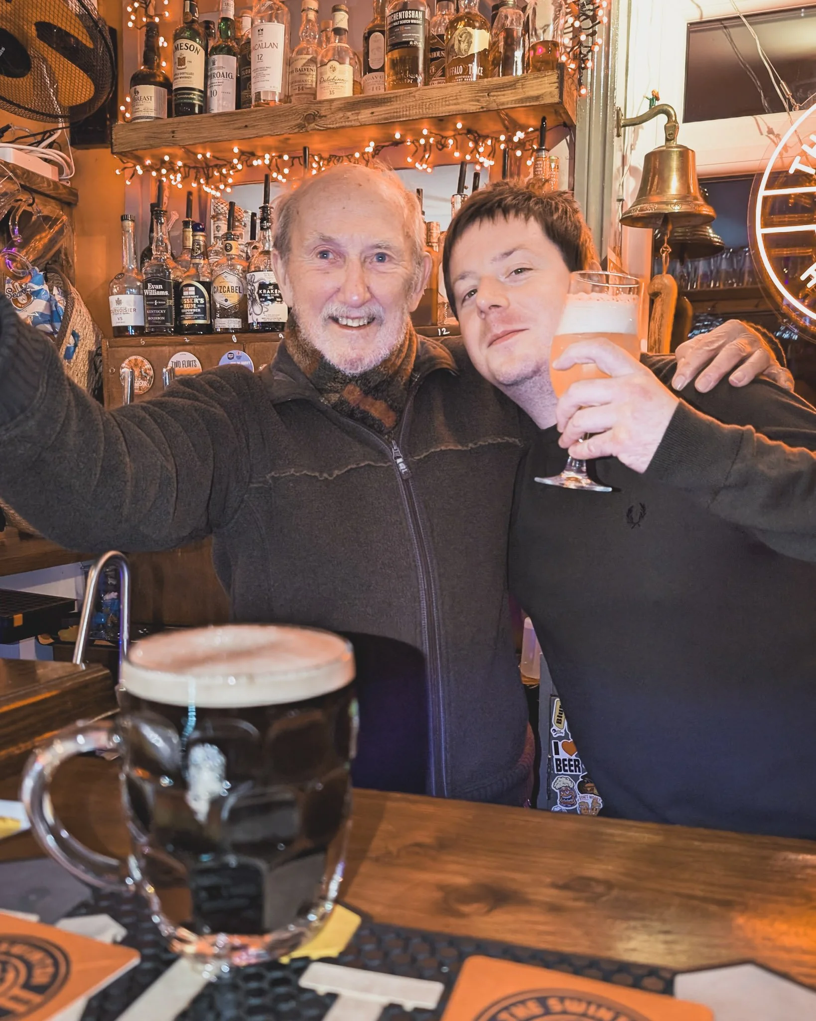 Two men at a bar taking a selfie, with one holding a beer and the other a glass of beer, smiling and enjoying drinks.