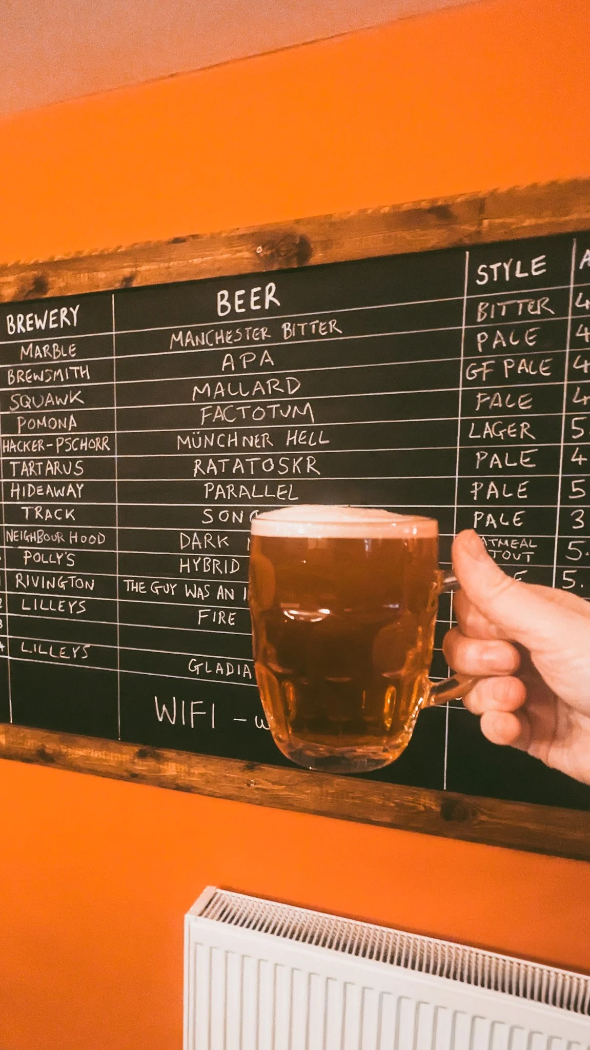 A hand holding a clear glass mug filled with beer in front of a blackboard menu listing different beers and their styles at a brewery or bar.