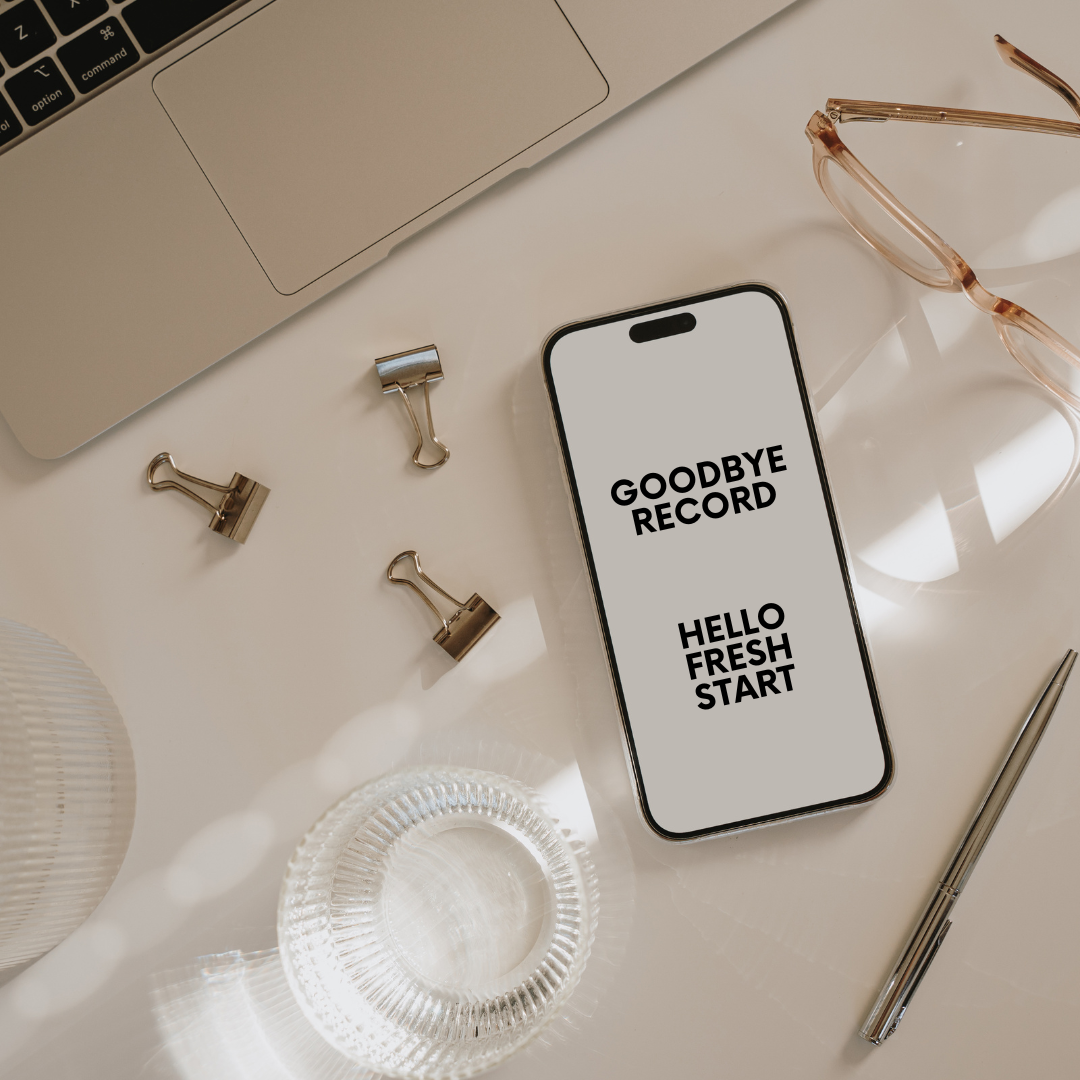 Smartphone on a desk with the text 'Goodbye Record Hello Fresh Start' on the screen. Surrounding items include glasses, a pen, paper clips, a glass dish, a laptop, and a white textured object.
