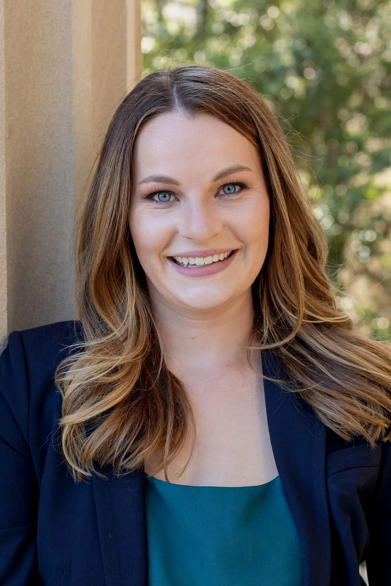 A young woman with blue eyes and long, wavy brown hair smiling outdoors, wearing a dark blazer and teal top, with blurred greenery in the background.