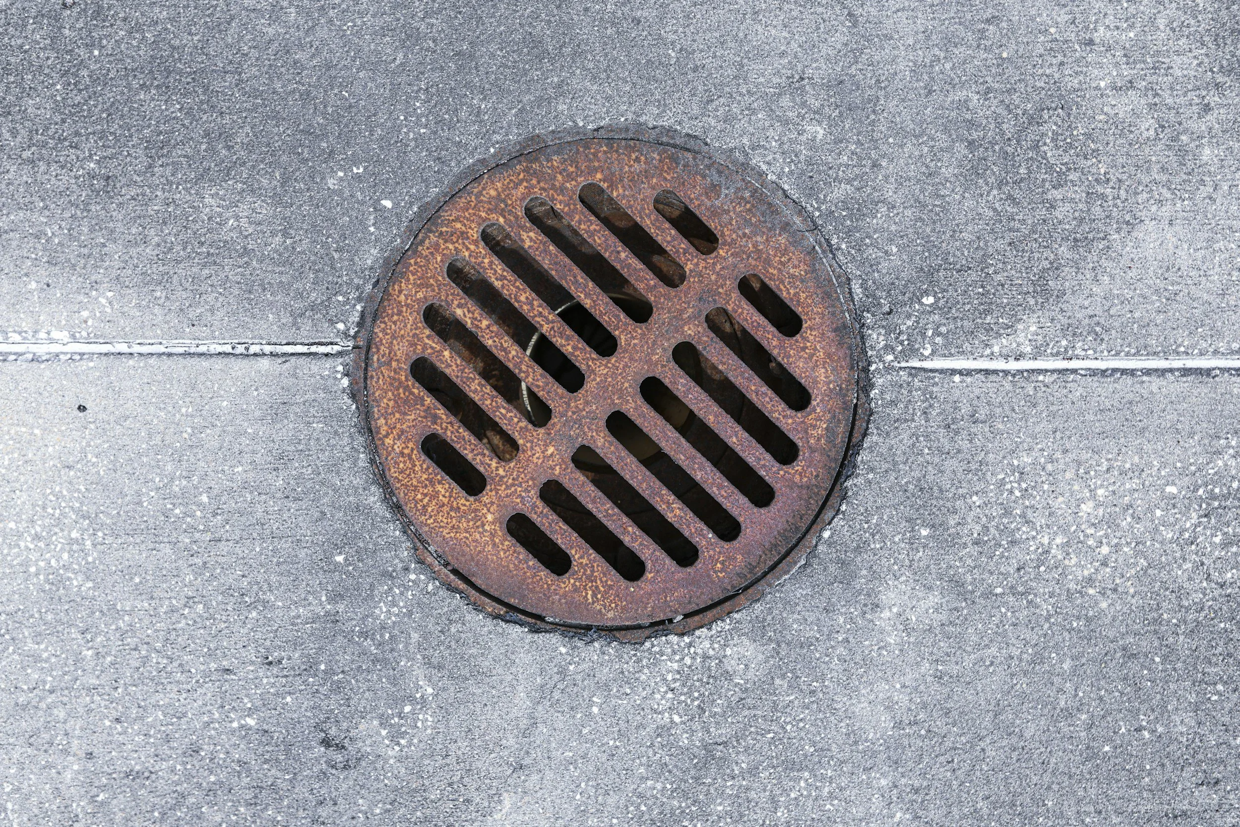 A circular metal storm drain cover on a concrete sidewalk, with rust and dirt around the edges.