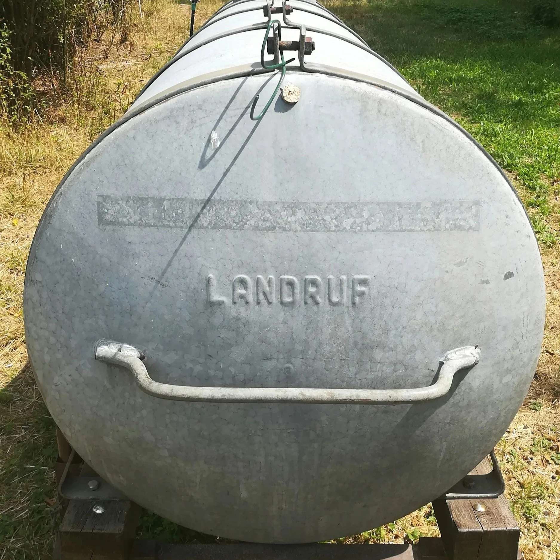 A large, round, metallic water or fuel tank with a handle and the word 'LANDRUF' embossed on the front, placed on a wooden pallet outdoors with grass and trees around.