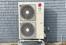 Outdoor air conditioning unit with two large fans, mounted on a metal stand against a gray brick wall.