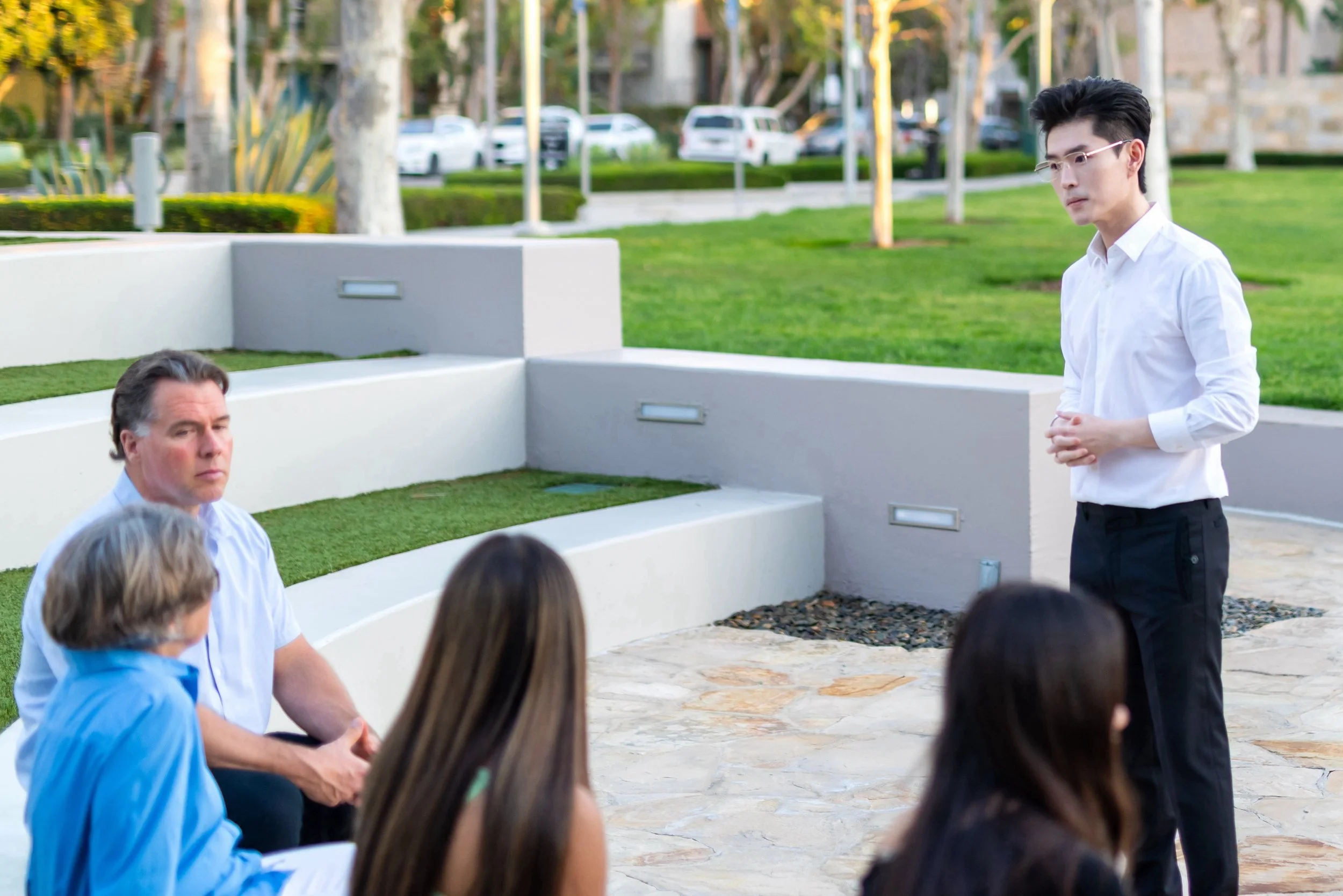A man wearing glasses and a white shirt standing outdoors in front of a group of seated people, with trees and parked cars in the background.