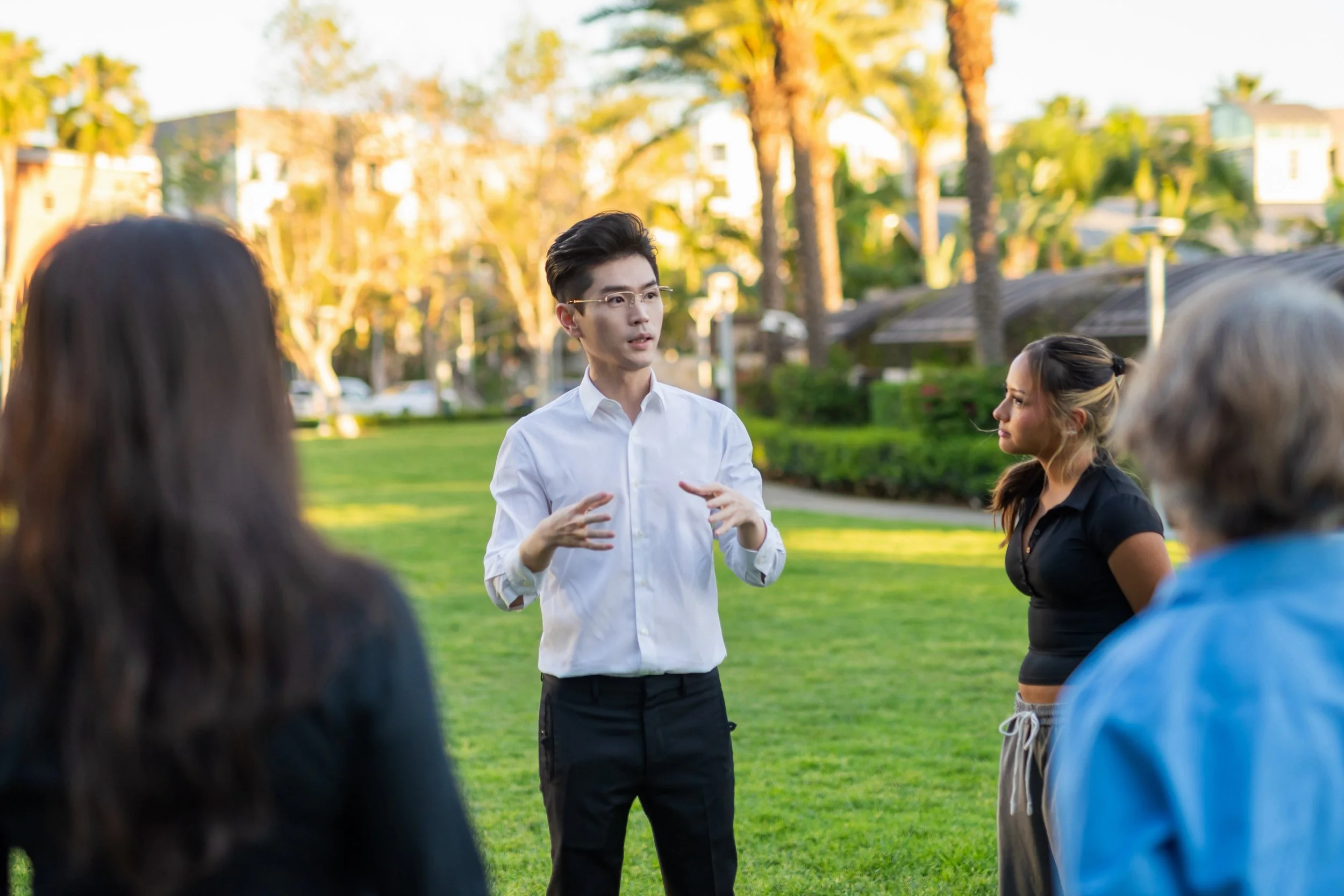 A man in a white shirt and glasses is speaking to a group of people outdoors in a park with green grass and trees during sunset. The group includes women of various ethnicities, listening attentively.