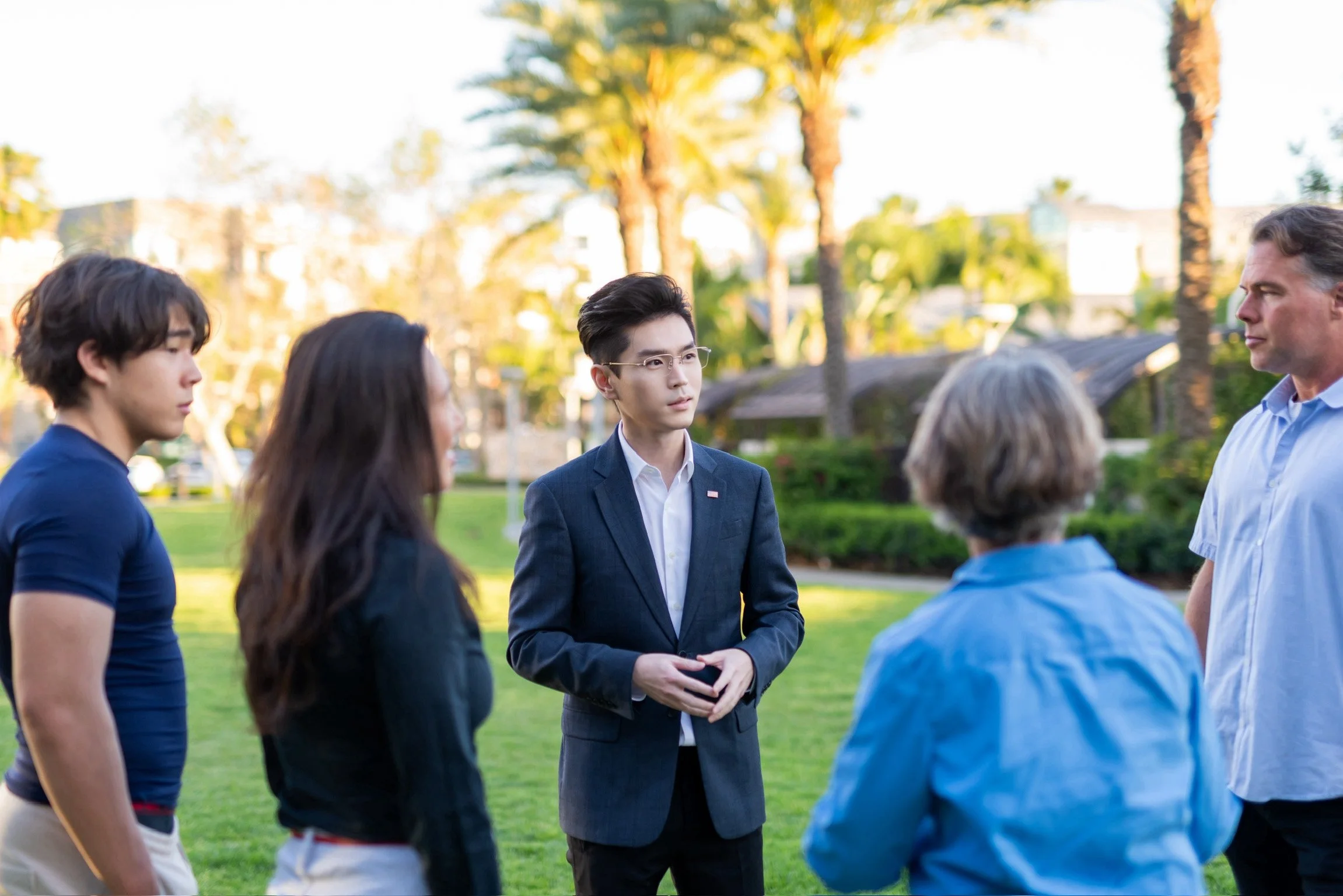 Group of five people standing outdoors on grass, engaged in conversation, with trees and houses in the background during daytime.