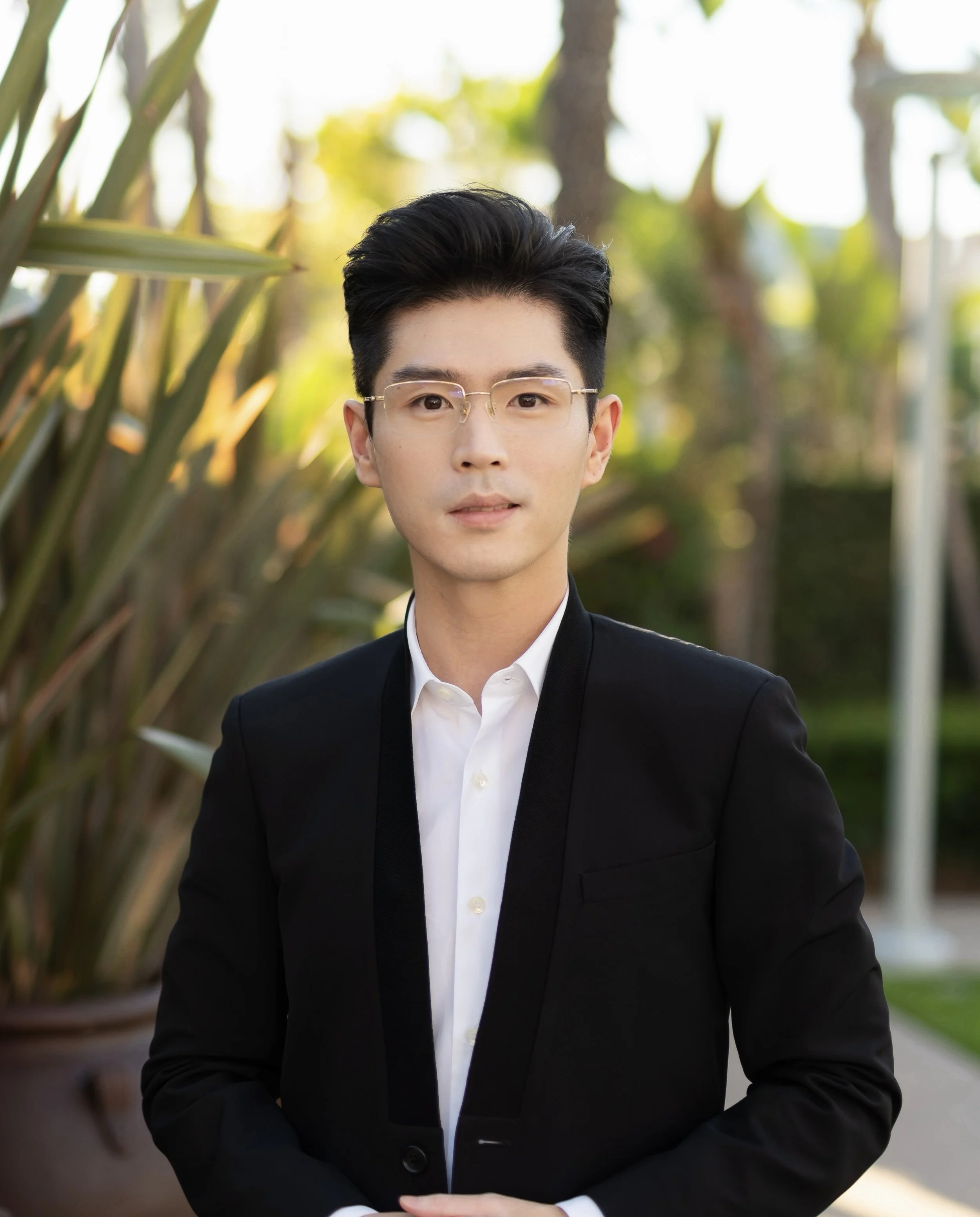 A young man with dark hair, wearing glasses, a white dress shirt, and a black suit, standing outdoors in front of greenery and trees with sunlight in the background.