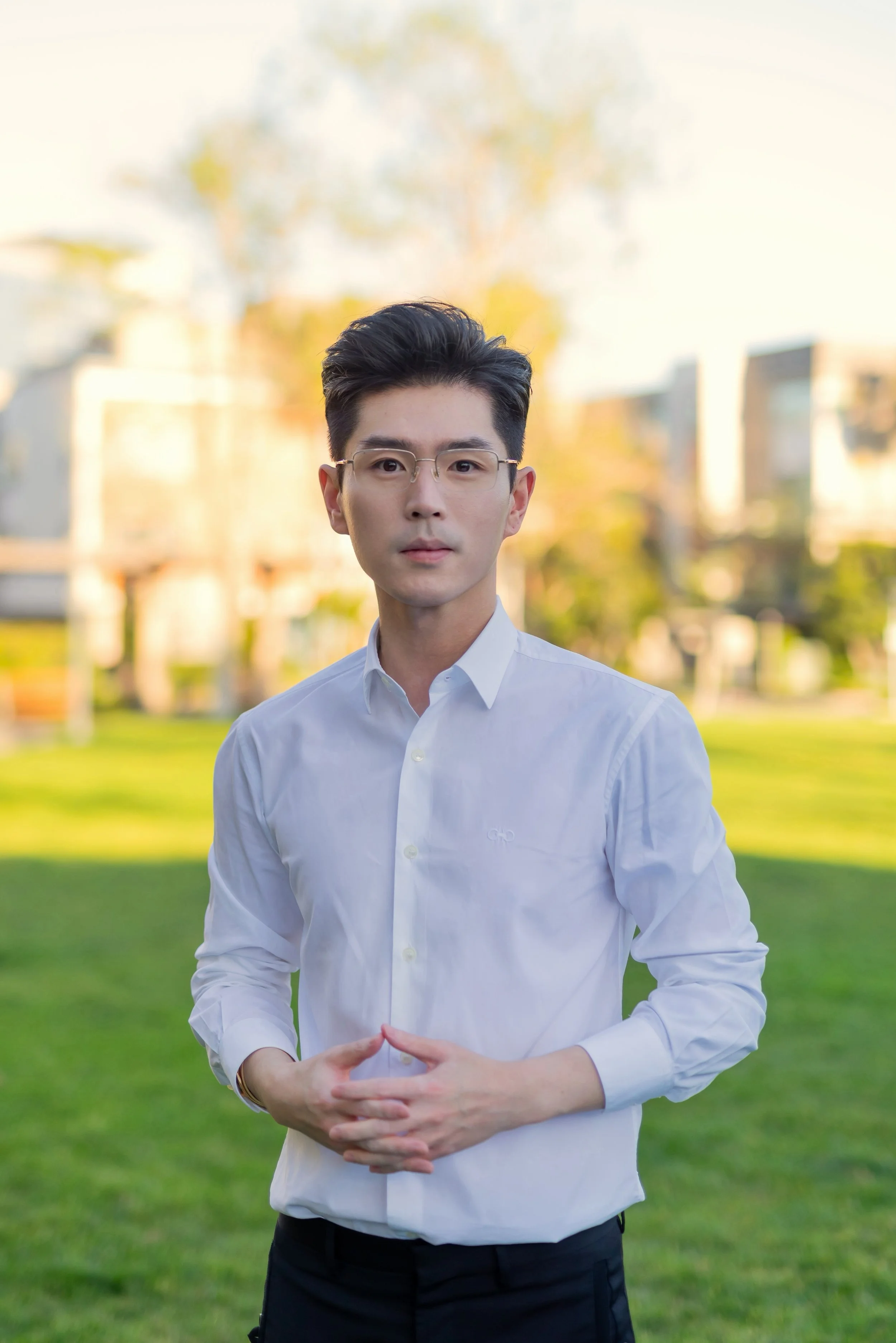 A young man with dark hair and glasses, wearing a white button-up shirt, standing outdoors with a blurred green park background during daytime.