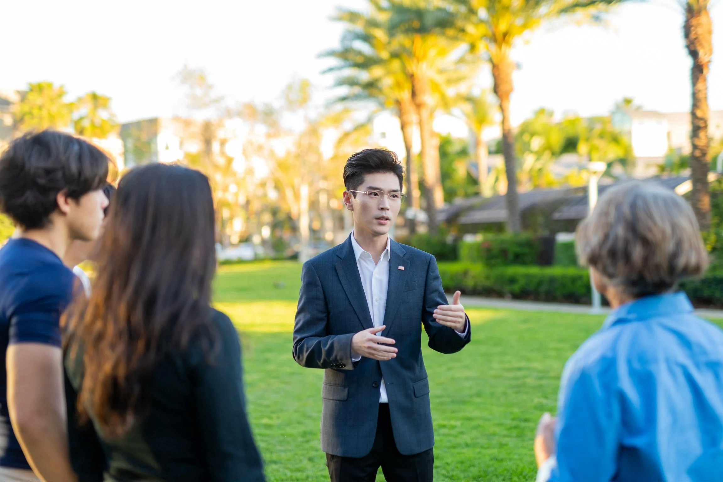 A group of people in conversation outdoors, with a man in a suit gesturing with his hands, in a park with palm trees and bright sunlight.