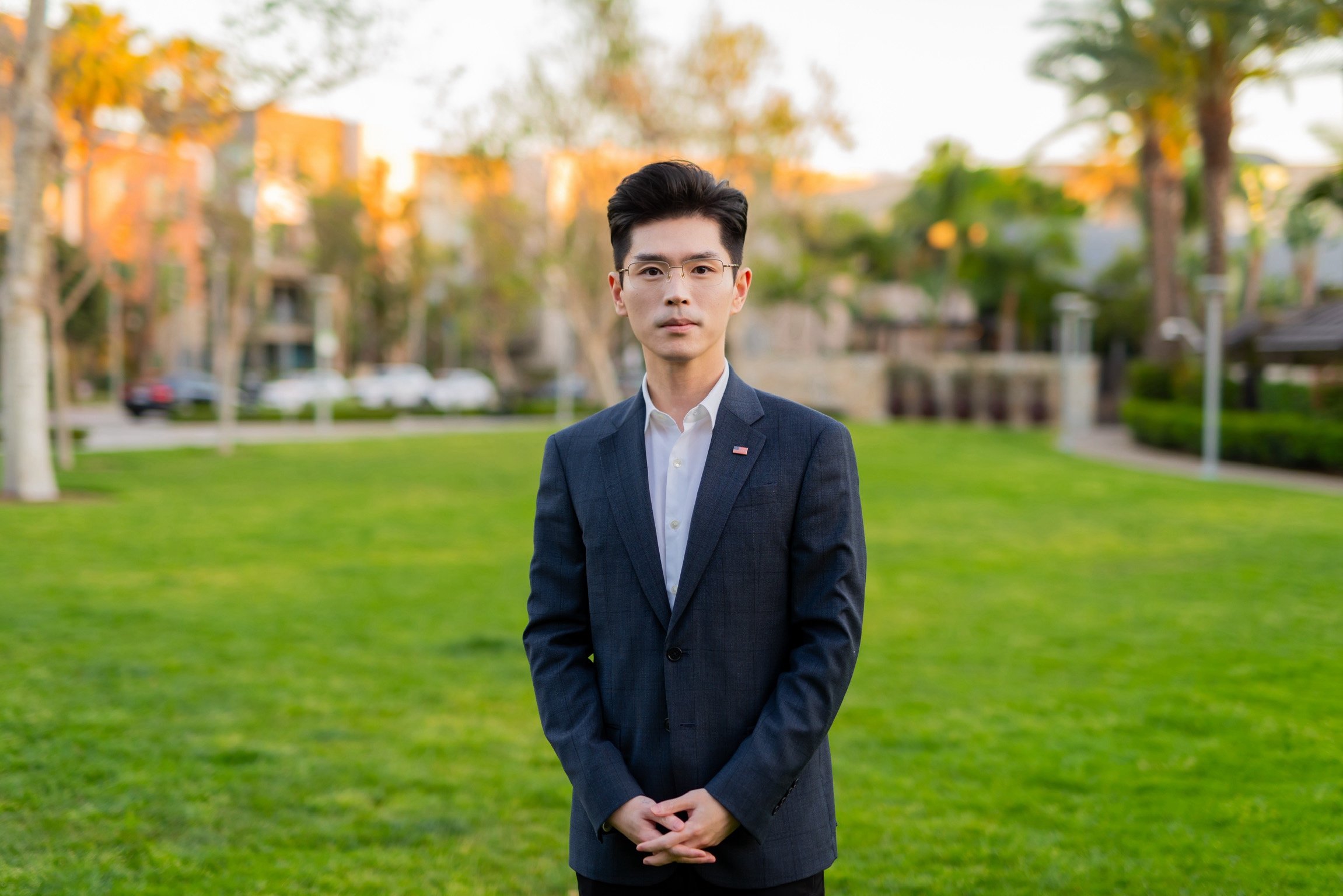 A man in a dark suit and white shirt standing outdoors on a grassy area with trees and buildings in the background during daylight.